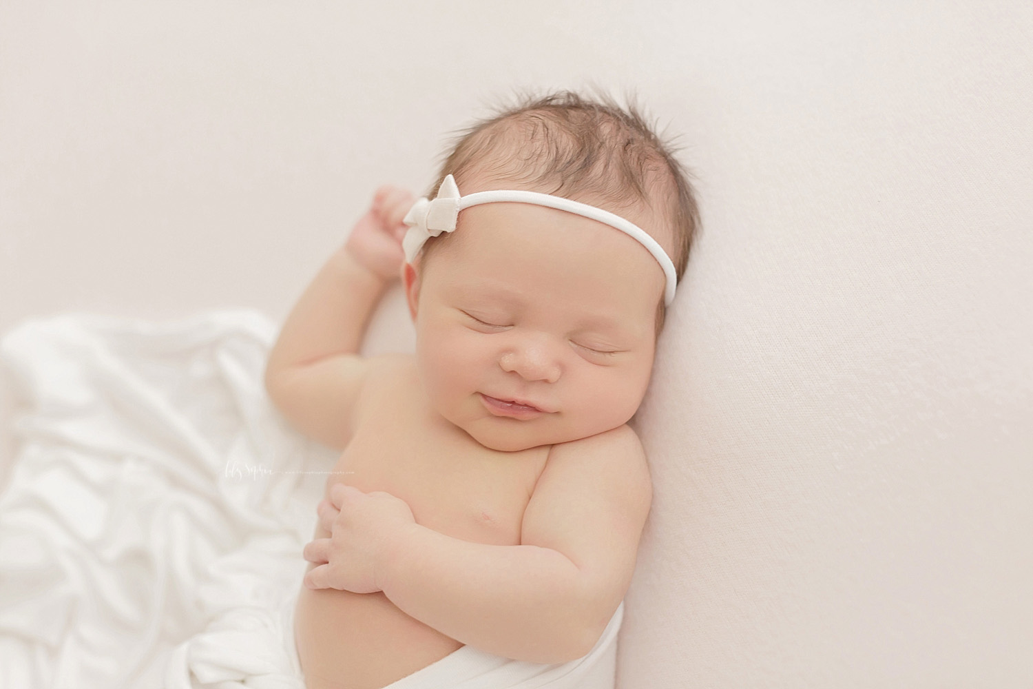  Image of a newborn, baby, girl, sleeping on her back, grinning, with one had on her tummy and one had by her head, wearing a white bow in her hair.&nbsp; 