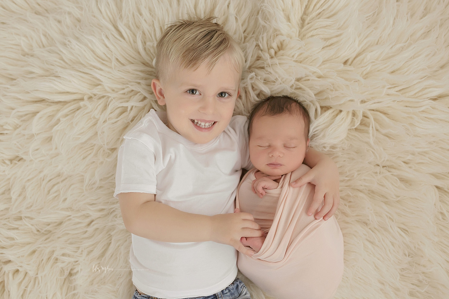  Image of a toddler boy wearing a white tee-shirt, smiling, laying beside his sleeping, baby, sister with his arm around her.&nbsp; 