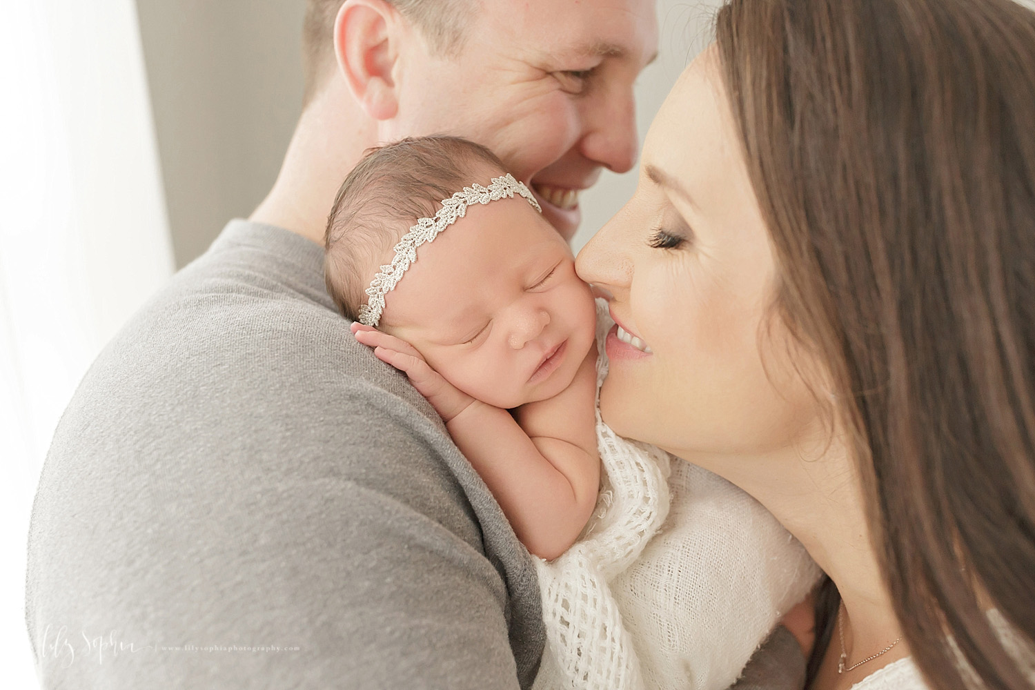  Image of a father, holding his sleeping, newborn, baby, girl on his shoulder while his wife kisses her daughter's cheek.&nbsp; 