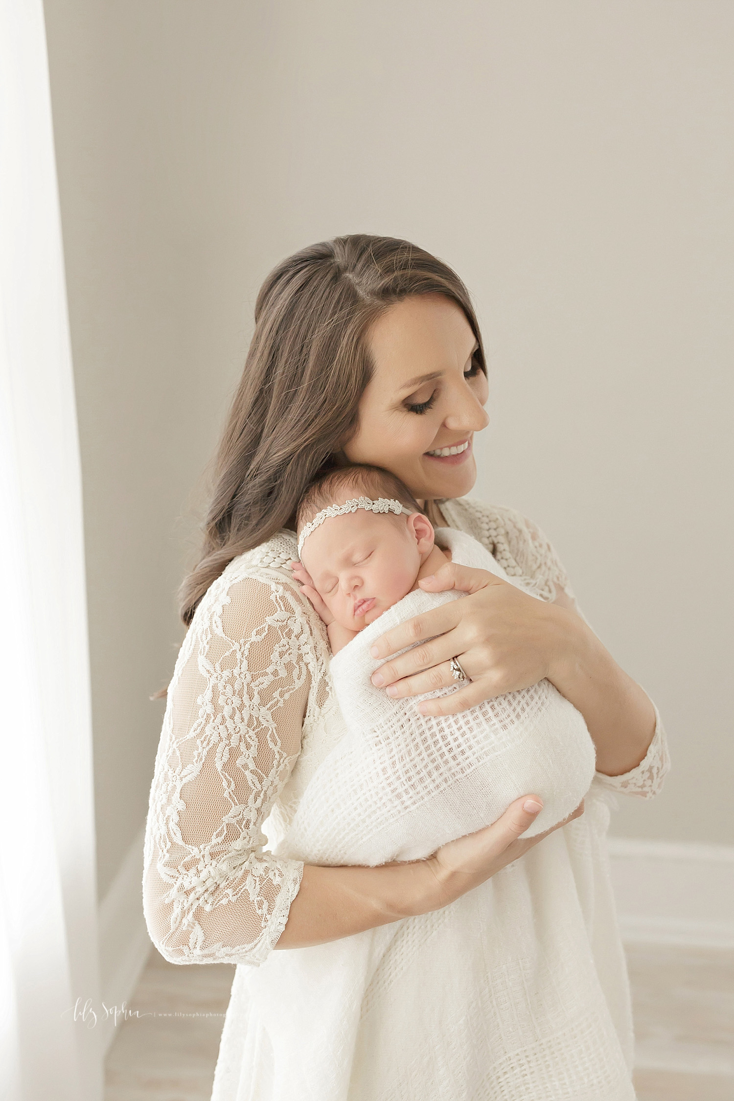  Image of a mother, wearing a lace top dress, holding her sleeping, newborn, baby, daughter, on her shoulder and smiling.&nbsp; 