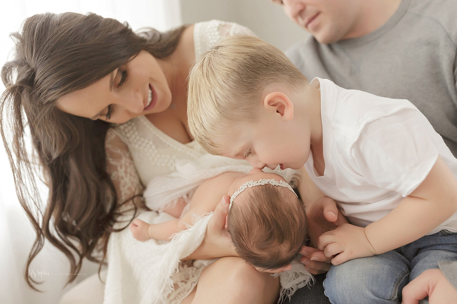  Image of parents, sitting on a couch, holding their newborn daughter on their lap while their 3 year old son leans over and touches his nose to his sister's. 