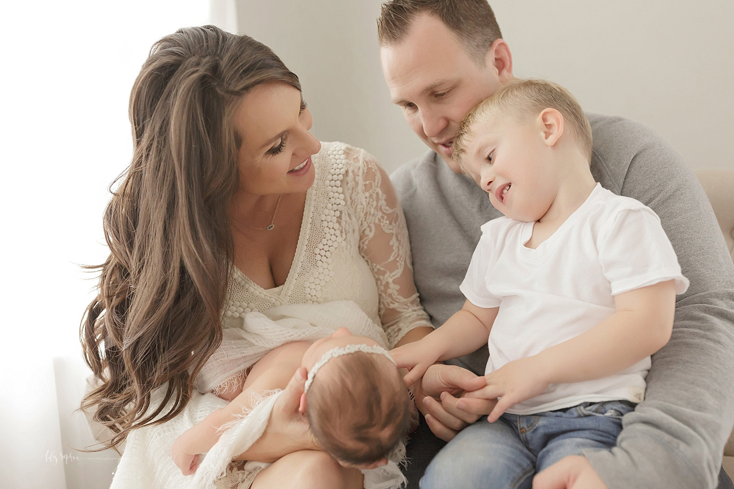  Image of a mother, sitting on a couch, holding her sleeping daughter on her lap while her husband holds their toddler son on his lap, and they all look down at the baby.&nbsp; 