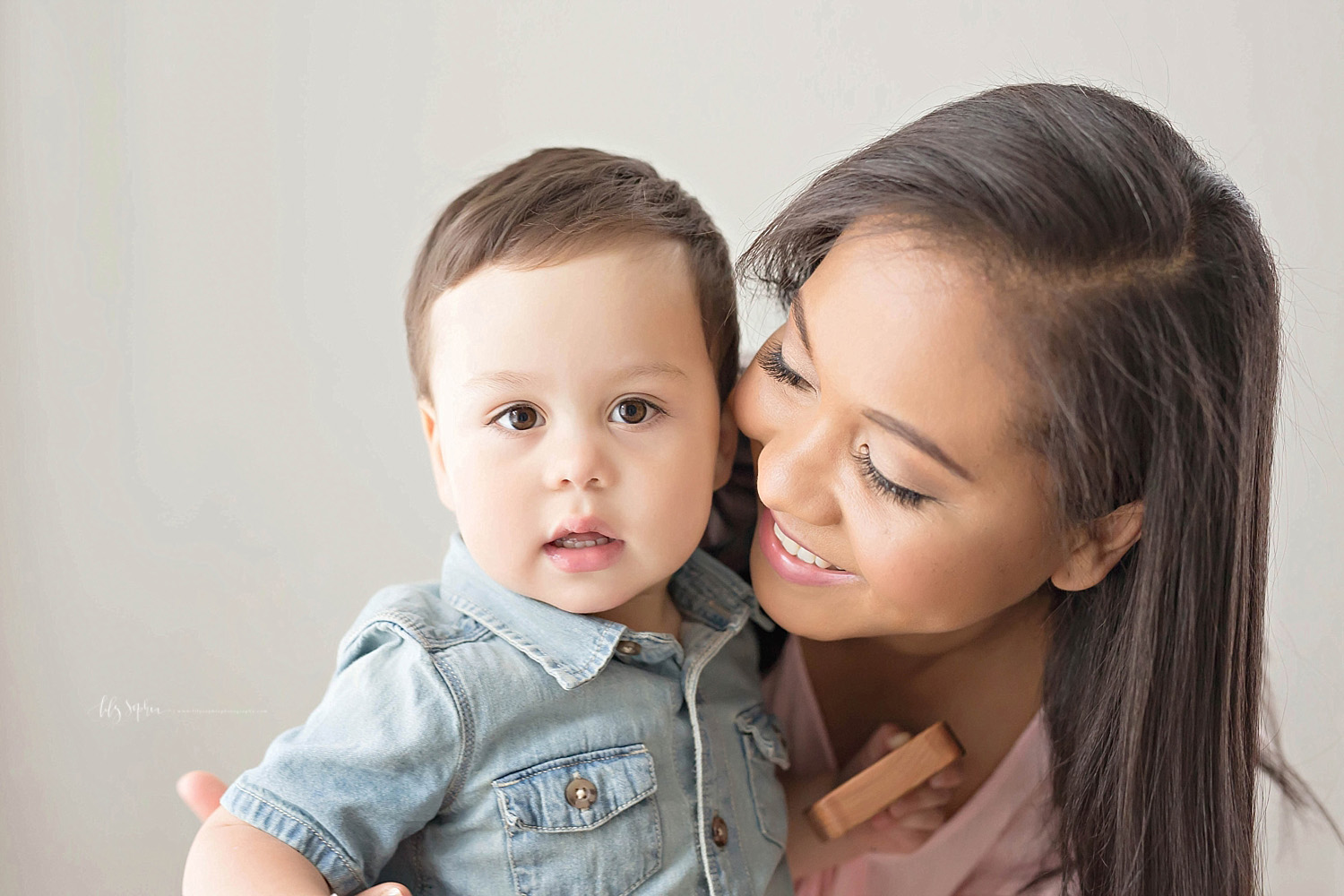  Image of a toddler boy, wearing a chambray shirt, looking at the camera with blocks in his hand, while his mother looks down at him and smiles.&nbsp; 