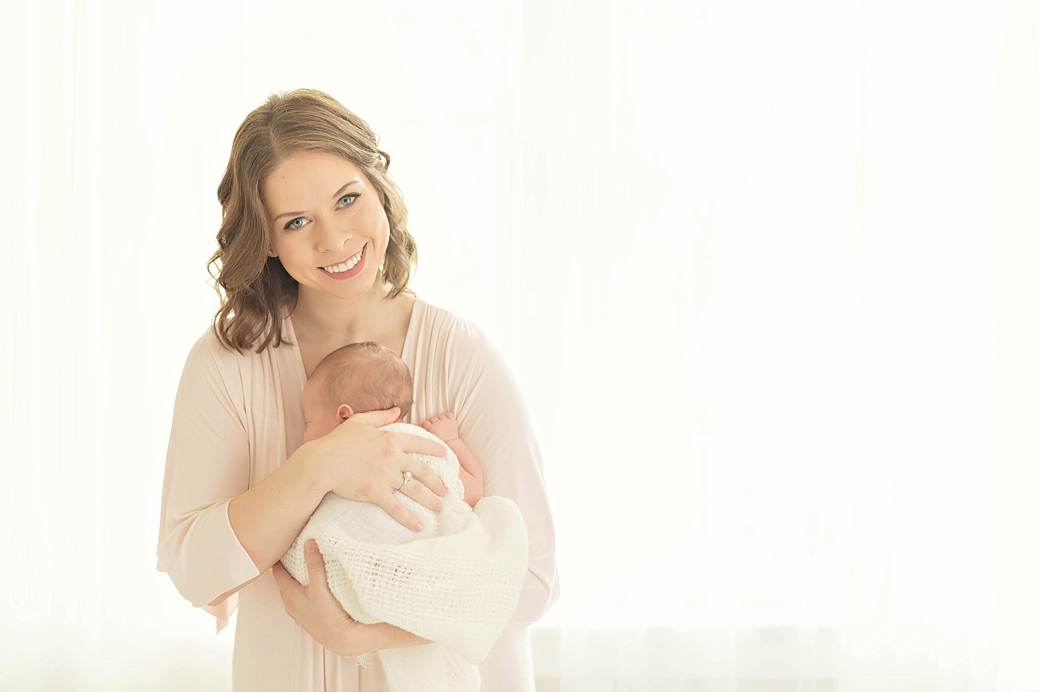  Image of a mother, standing in front of a window, smiling as she cradles her sleeping, newborn, son to her chest.&nbsp; 