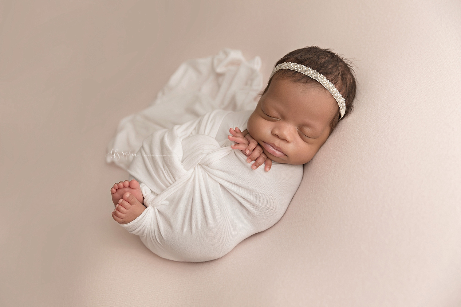  Image of an African American baby girl, wrapped with a white wrap with just her fingers and toes peeking out and a pearl tie back in her hair.  