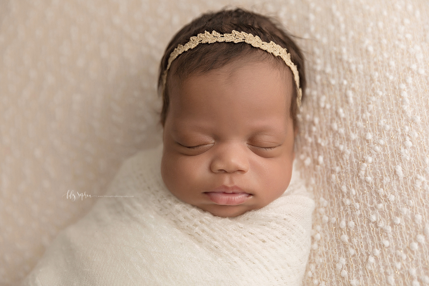  Image of a sleeping, African American, baby, girl, with a gold leaf tie back in her hair, wrapped up tight in a cream wrap.  