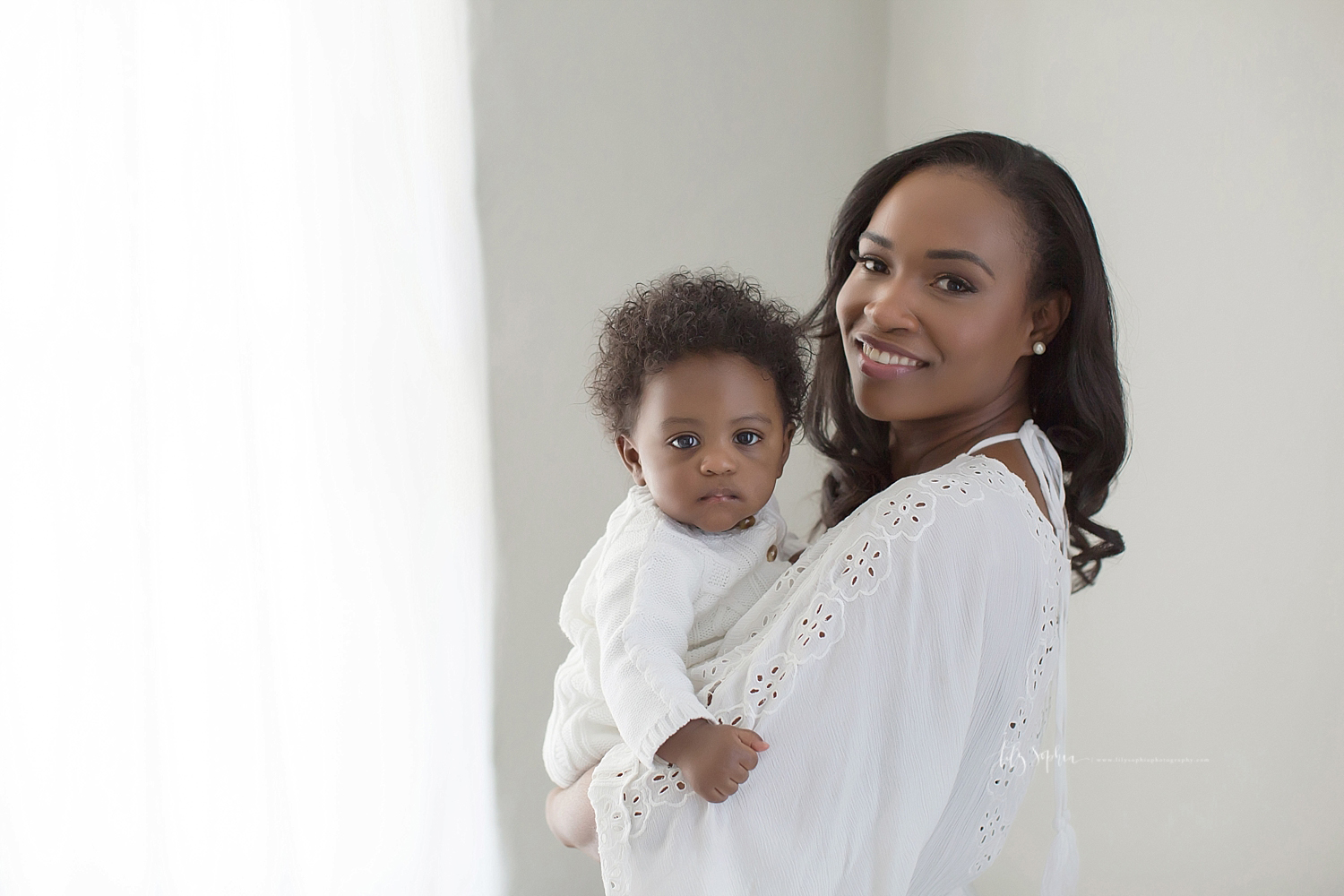  Image of an African American woman, wearing a while lace dress, holding her 6 month old son who is wearing a white romper.  