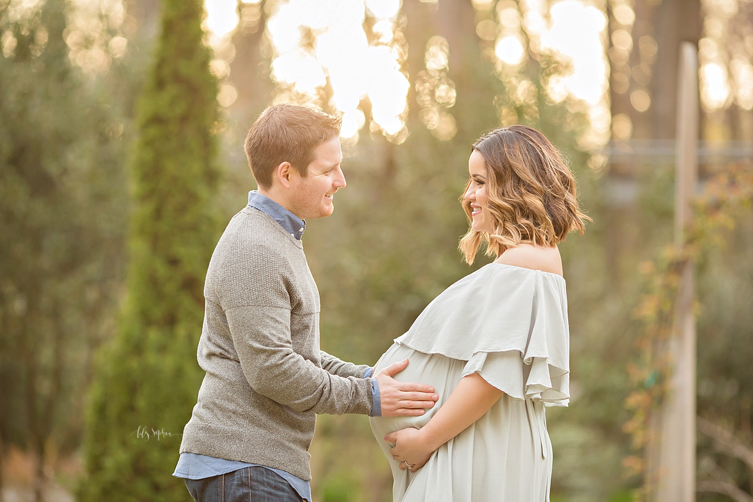  Image of a pregnant woman, and her husband, facing each other and standing in a garden, at sunset. 