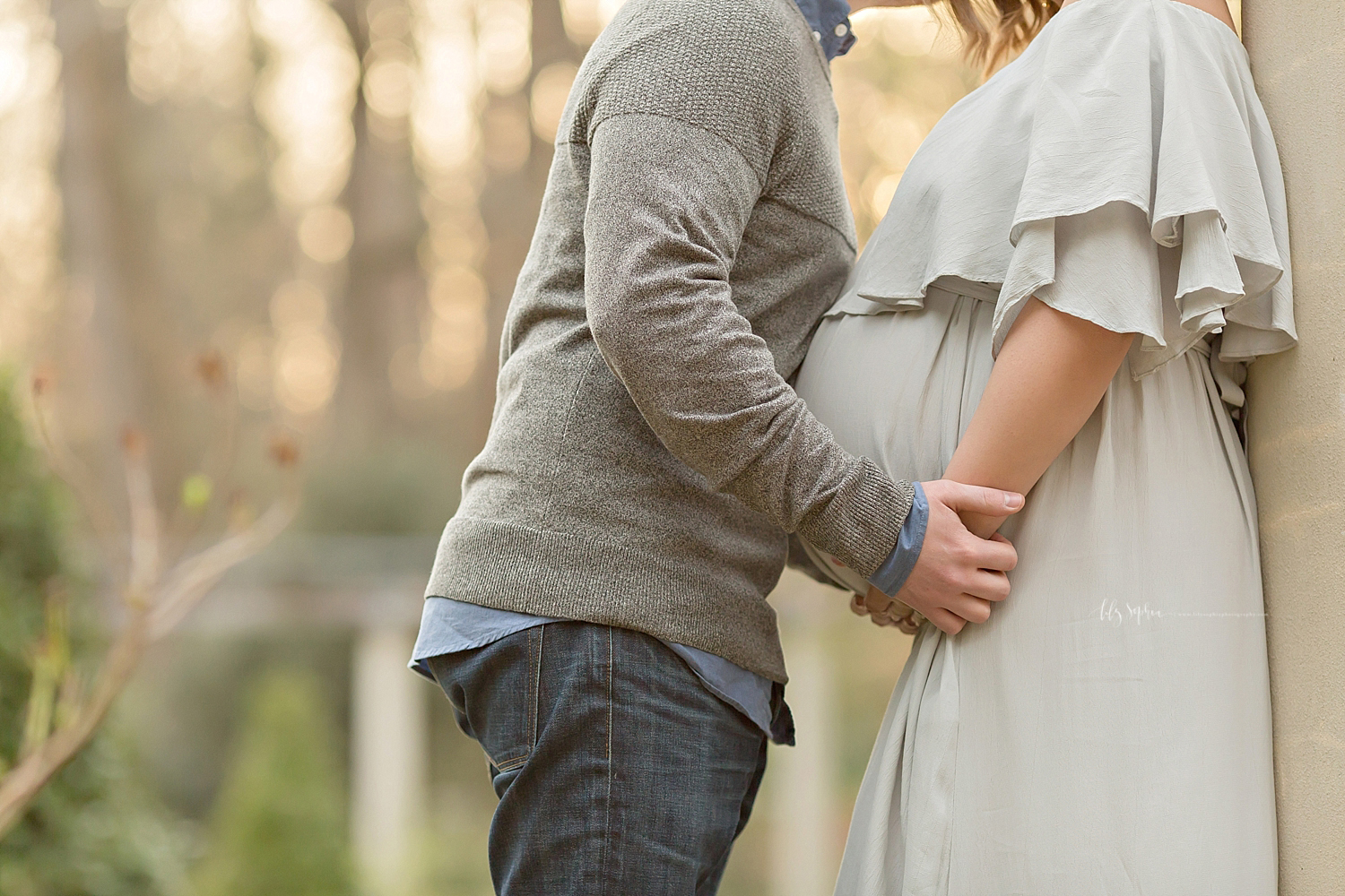 Image of a pregnant woman, and her husband, standing in a garden, with her hands under her belly as he holds her wrists  