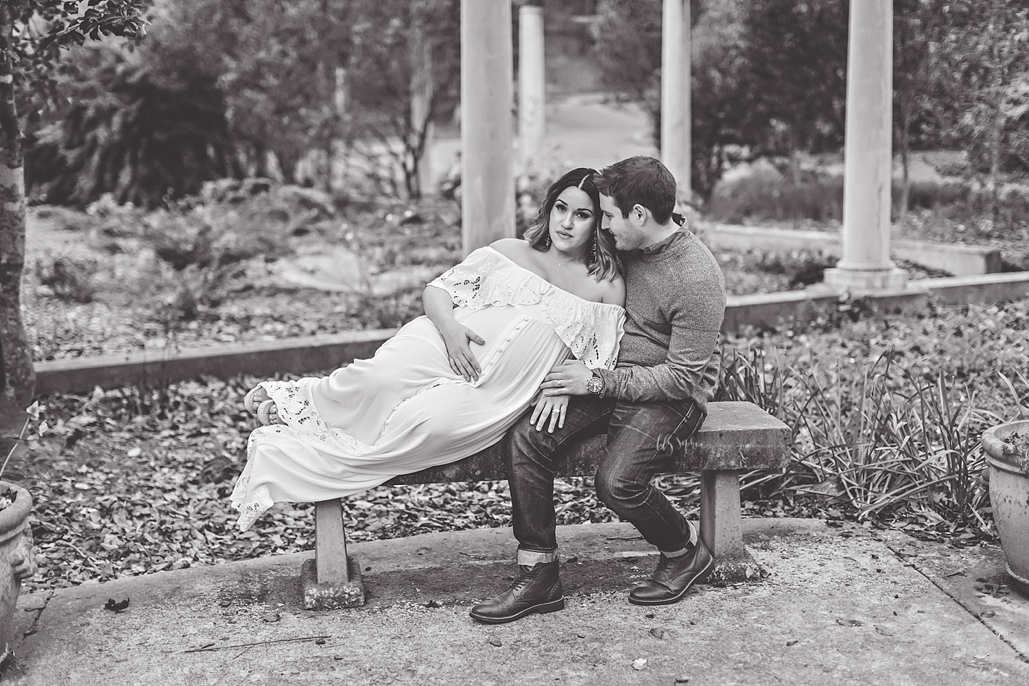  Black and white image of a man sitting on a stone bench, smiling down at his pregnant wife leans on him and has one hand on her belly.  