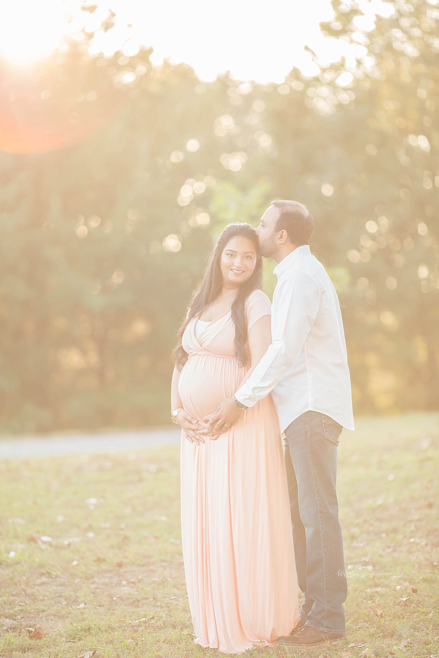 Image of a pregnant, Indian, woman and her husband, standing in a park at sunset, while he kisses her head, while she smiles. 