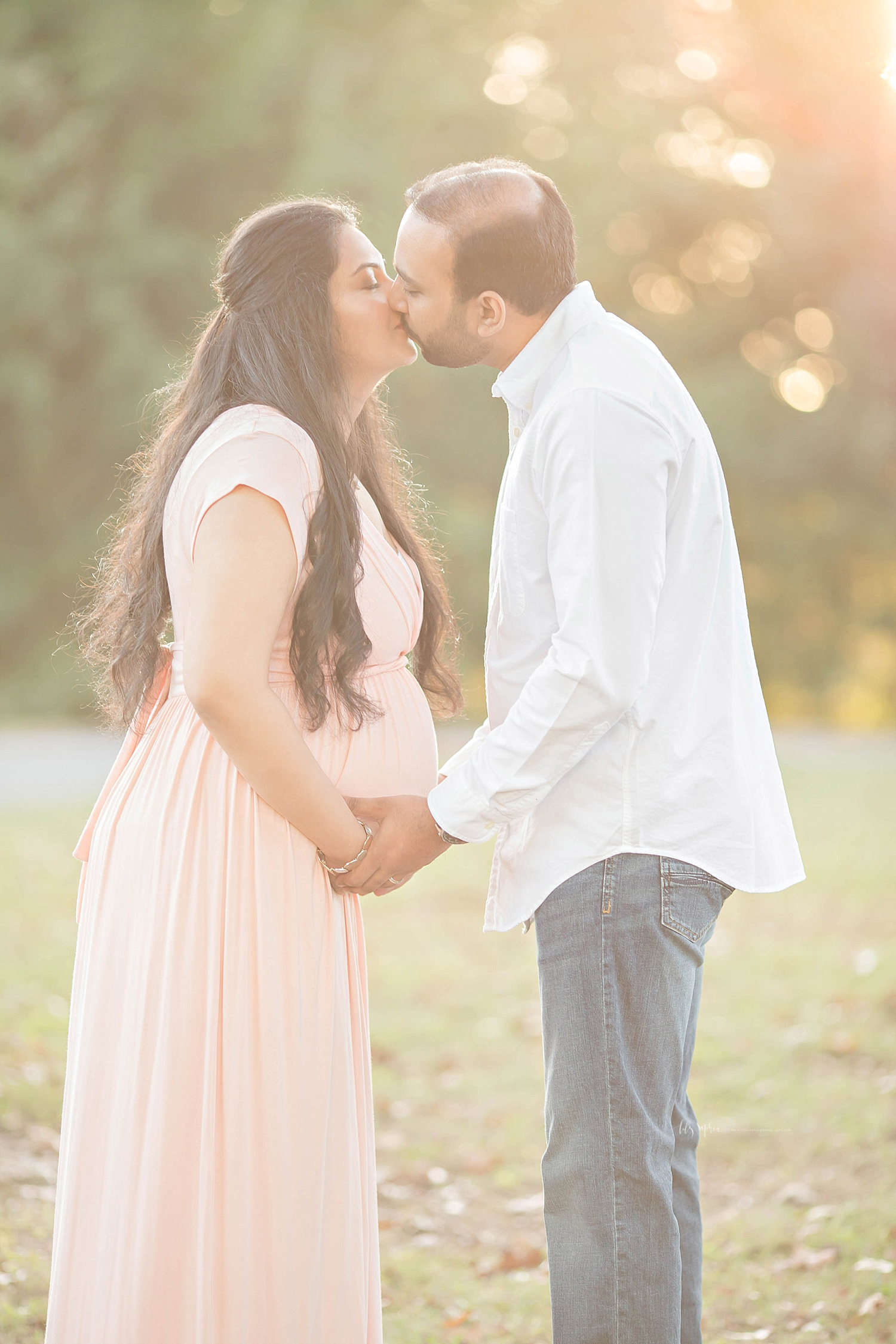 Image of a pregnant couple, holding hands and kissing in a backlit park. 