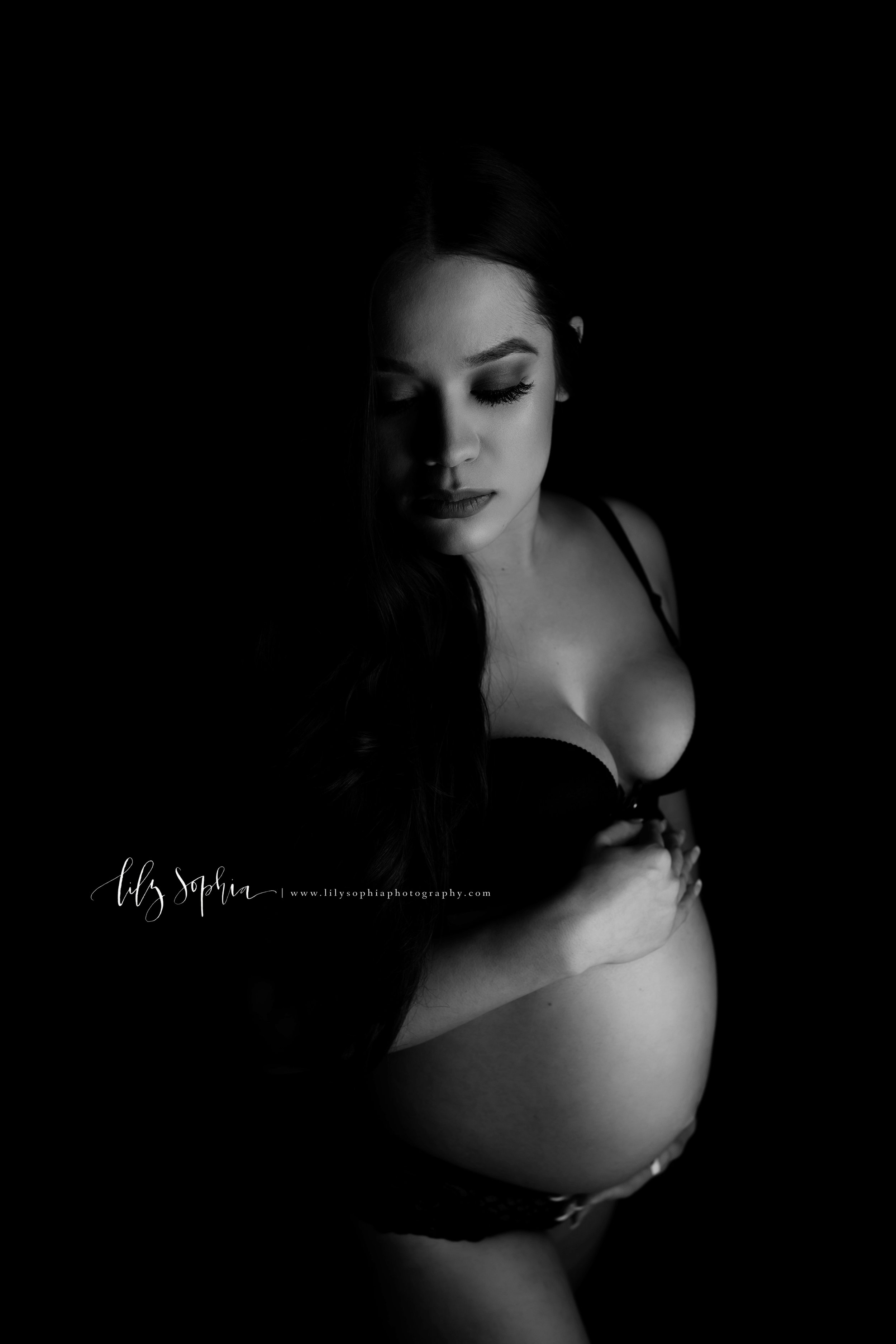  Black and white editorial maternity fine art image of beautiful pregnant Hispanic woman with long dark hair dressed in black lace bra and robe  with a black background in the Atlanta studio of Lily Sophia Photography.  