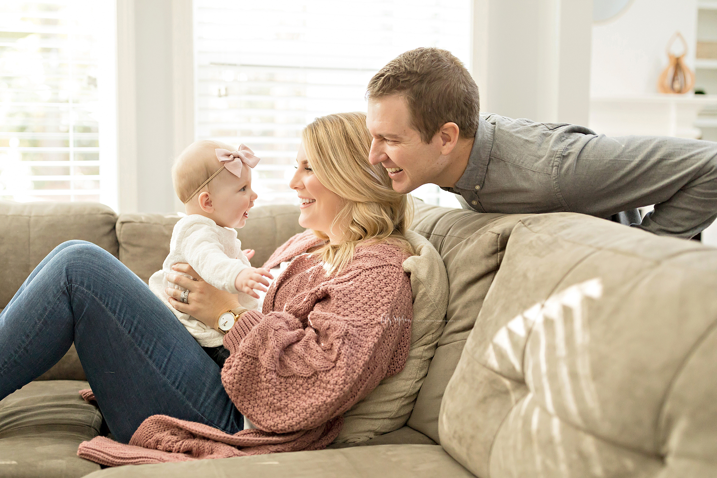  Photograph of a happy family of three in their home in natural light.. Mom is sitting on a tan sofa with her knees bent. She is holding her baby girl on her lap. Dad is behind the sofa peering over Mom’s left shoulder. Blonde haired Mom is wearing a