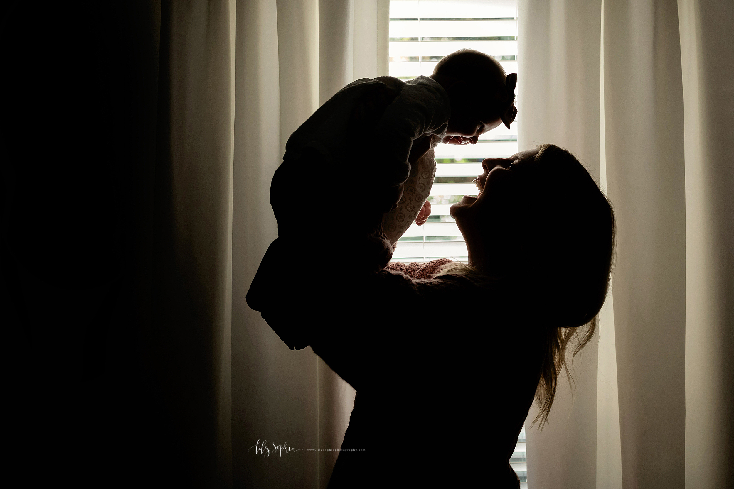  Silouhette profile image of a mom and her baby girl in front of a window in their home.  Mom is standing holding her daughter under her arms and lifting her above her head.  Both Mom and the daughter are smiling at one another as the light filters i