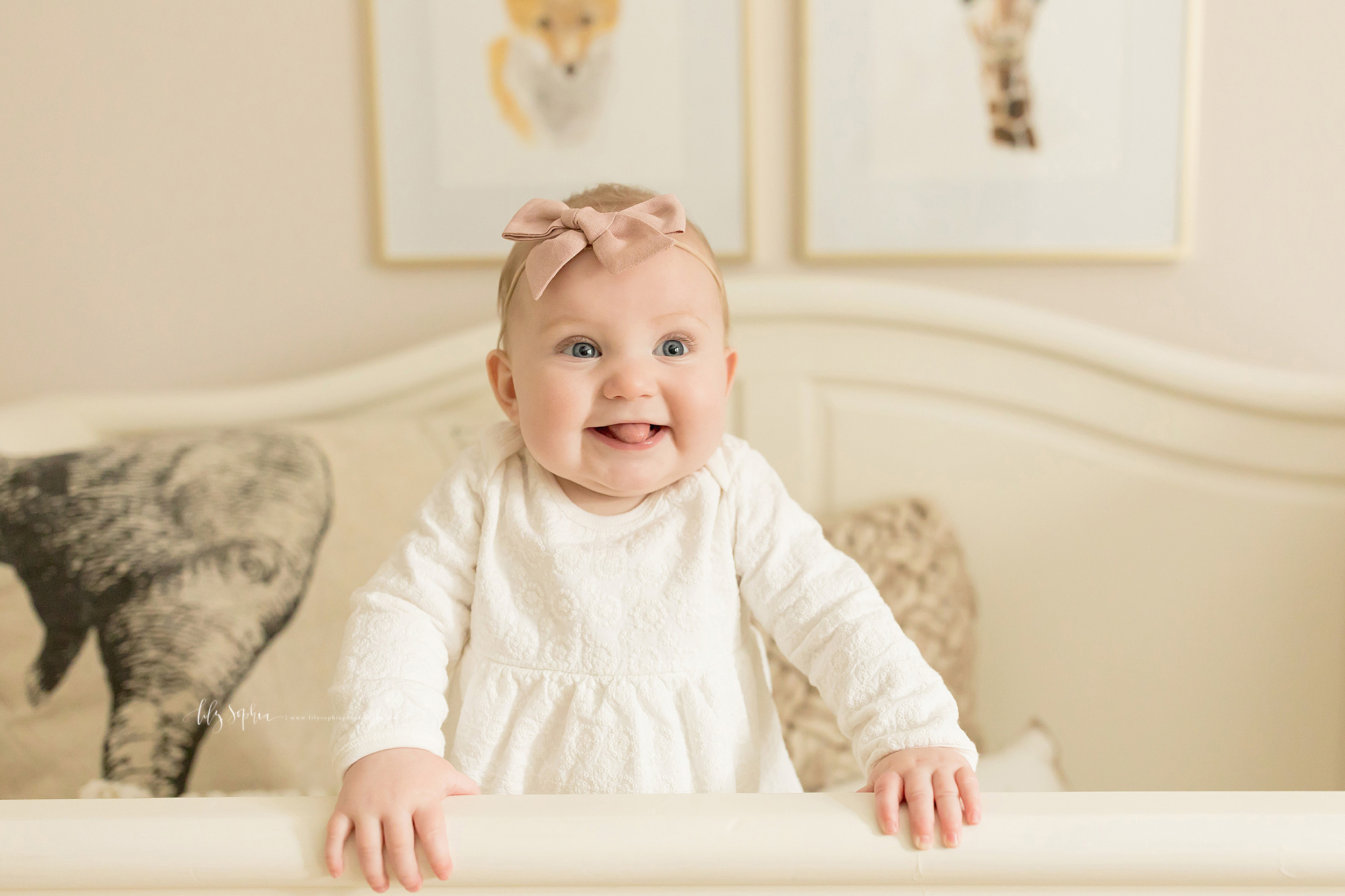  Photograph of a baby girl in natural light in her nursery.  The blonde haired, blue-eyed baby girl is standing in her crib and holding the crib rail.  She is bright-eyed and smiling with her tongue sticking out of her mouth. 