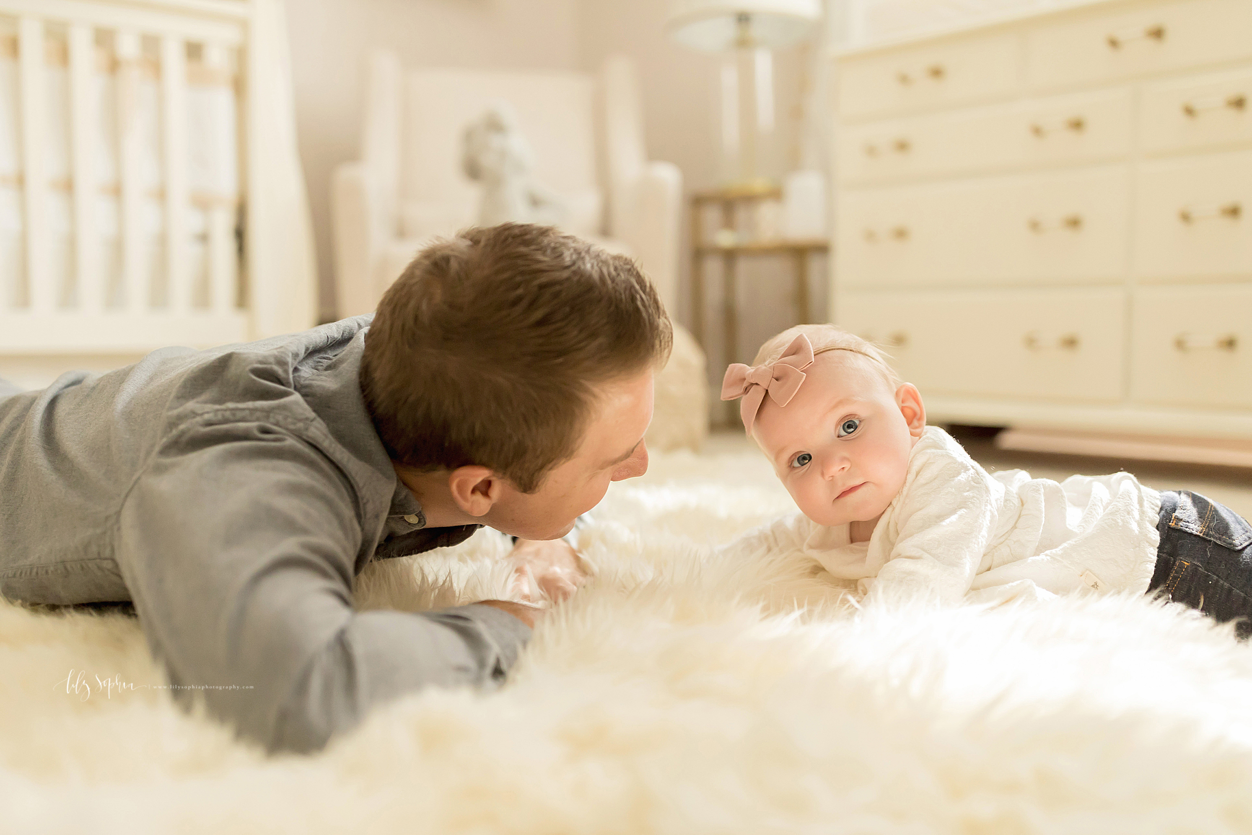  Image of dad and blonde haired blue-eyed baby daughter on a white furry rug in the nursery of their home. Dad is wearing a grey long sleeve buttoned down shirt and is laying on his stomach with his arms folded and lifting his head and chest off the 