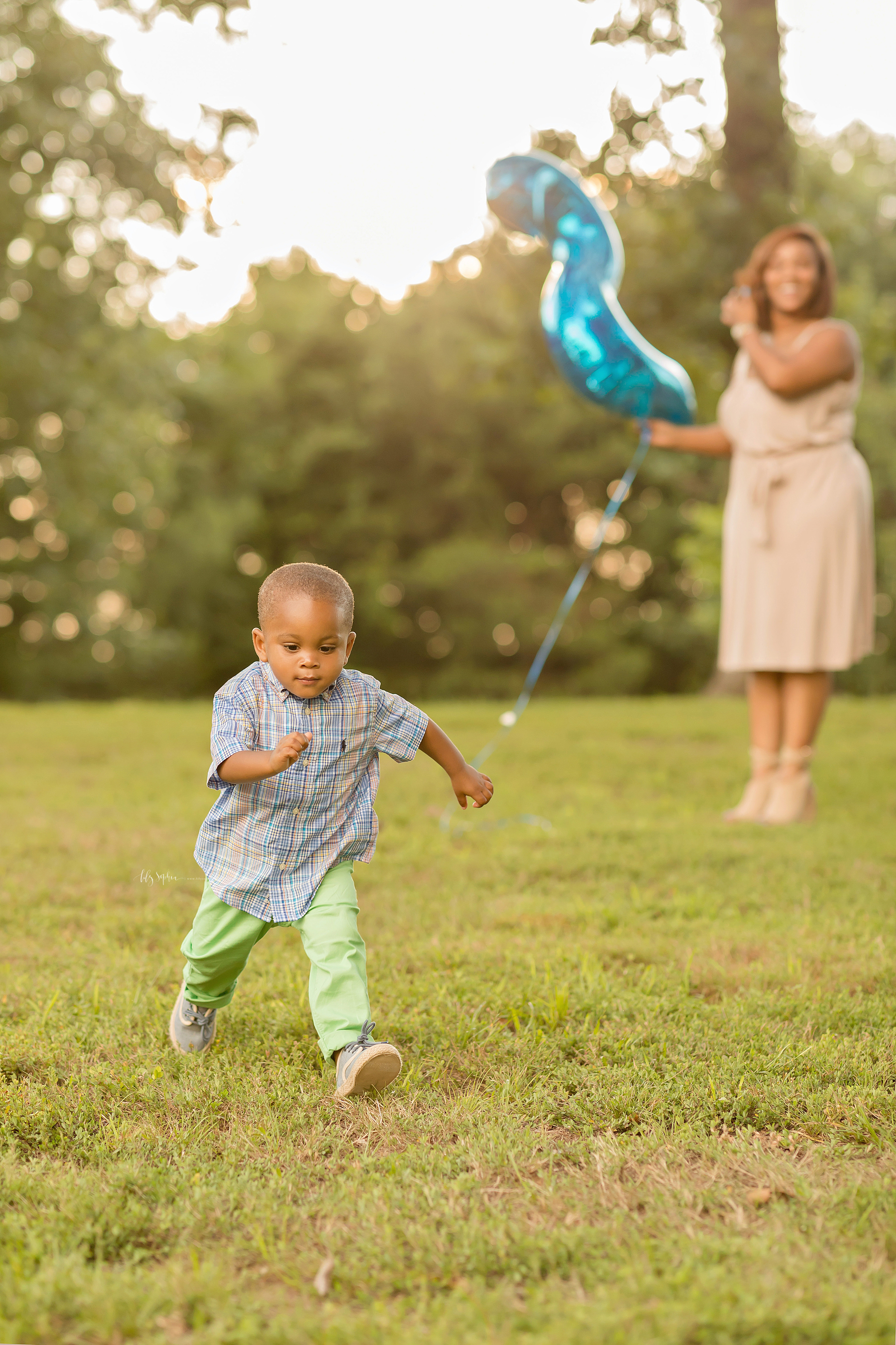  African American two year old with big brown eyes running through the park on his second birthday while his mom holds his blue number two balloon in the background. 