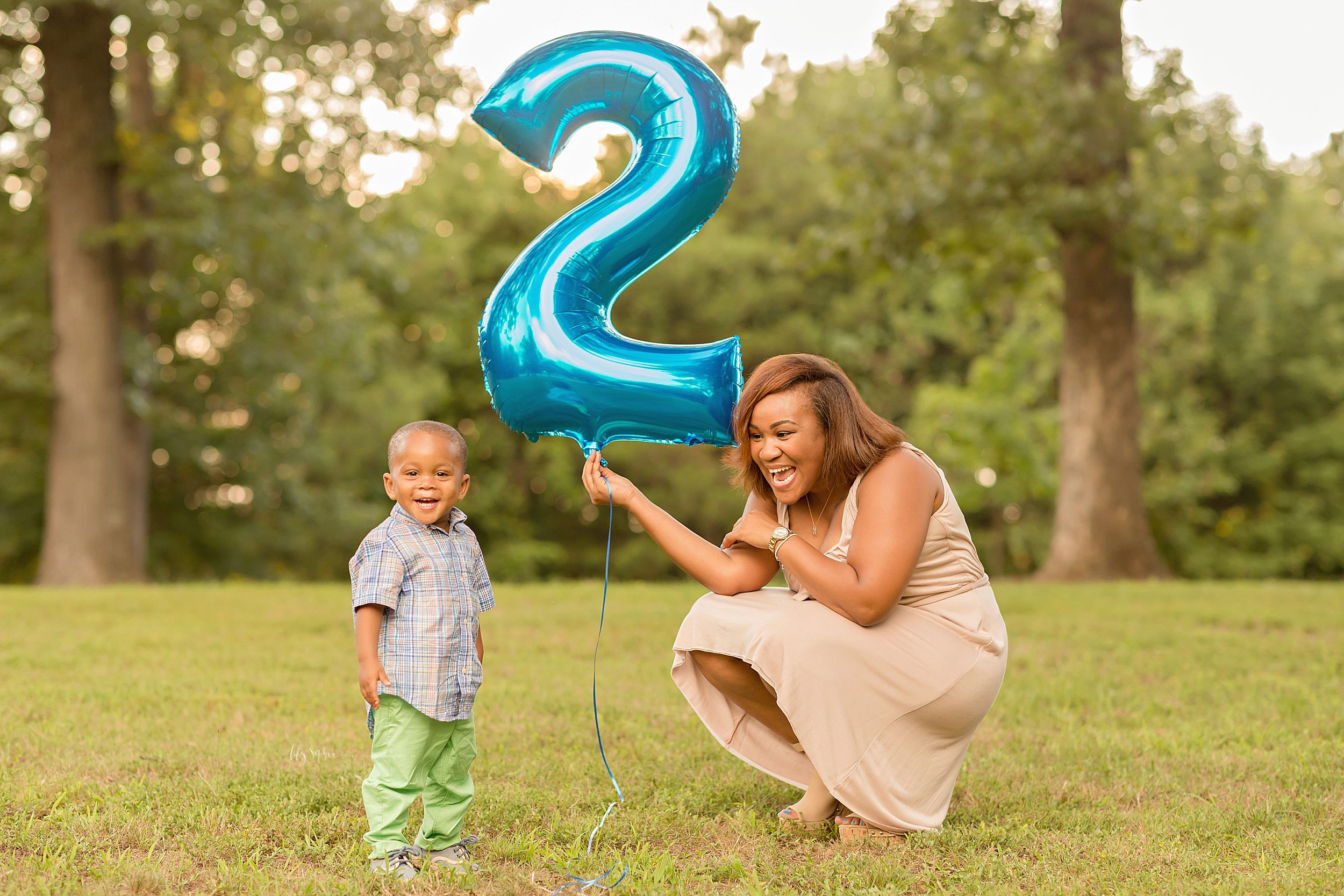  Photo of an African American mom and her son celebrating his two year old birthday in an Atlanta park.  Mom is squatting down in a taupe sleeveless dress holding a blue number two balloon.  Her son is standing next to her in a blue and tan plaid but