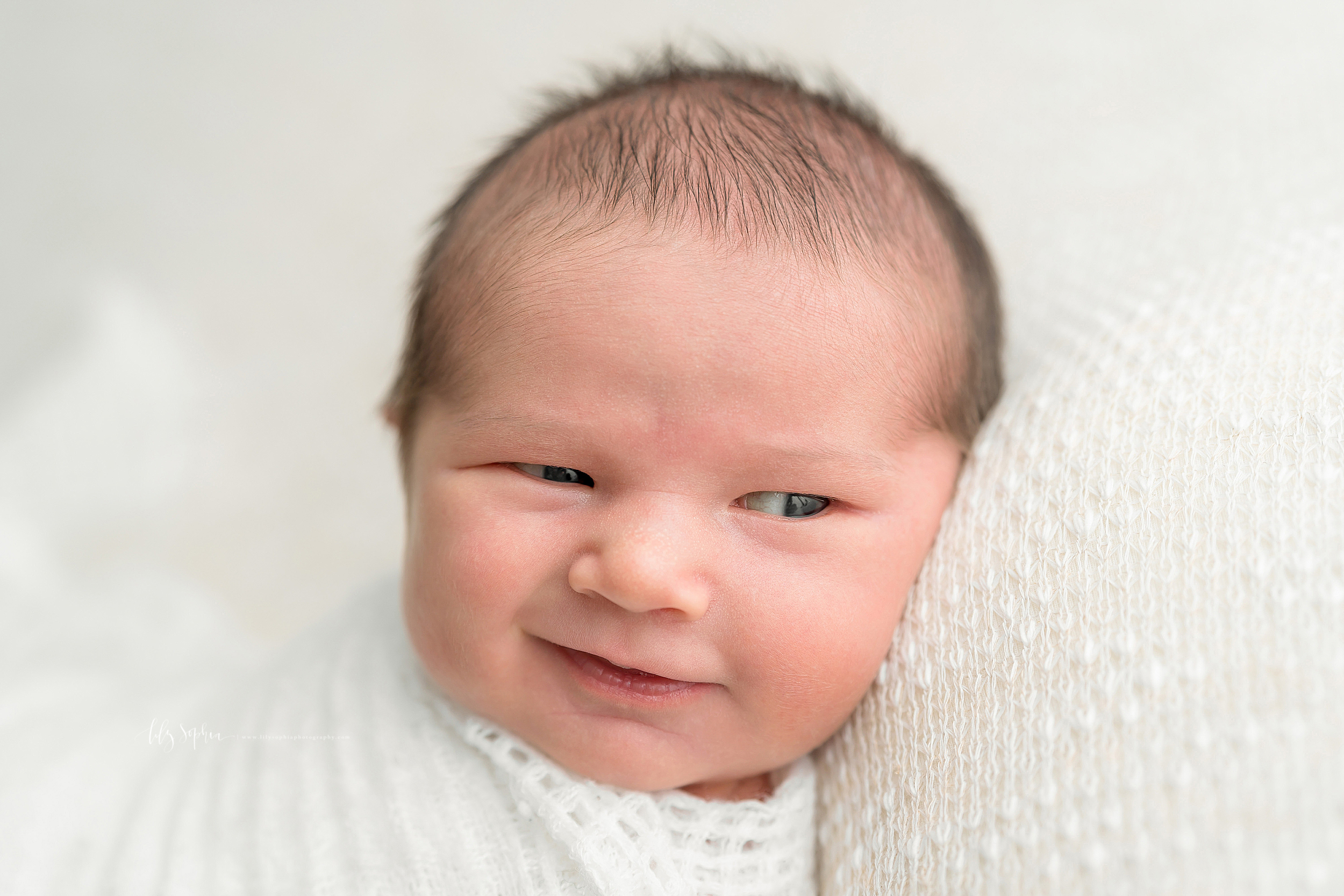  Atlanta newborn baby boy with wispy brown hair wrapped in a white blanket.  He is grinning with his eyes sparkling. 