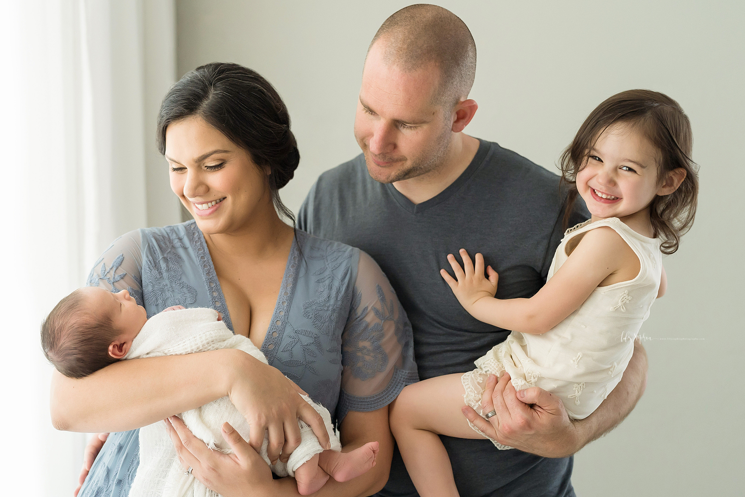  Family of four are taking photographs at Lily Sophia Photography Studio in Atlanta.  Mom, a brunette, is wearing a blue lace dress while holding her infant son in her arms and smiling at him.  Dad is behind Mom holding his toddler daughter who is ex