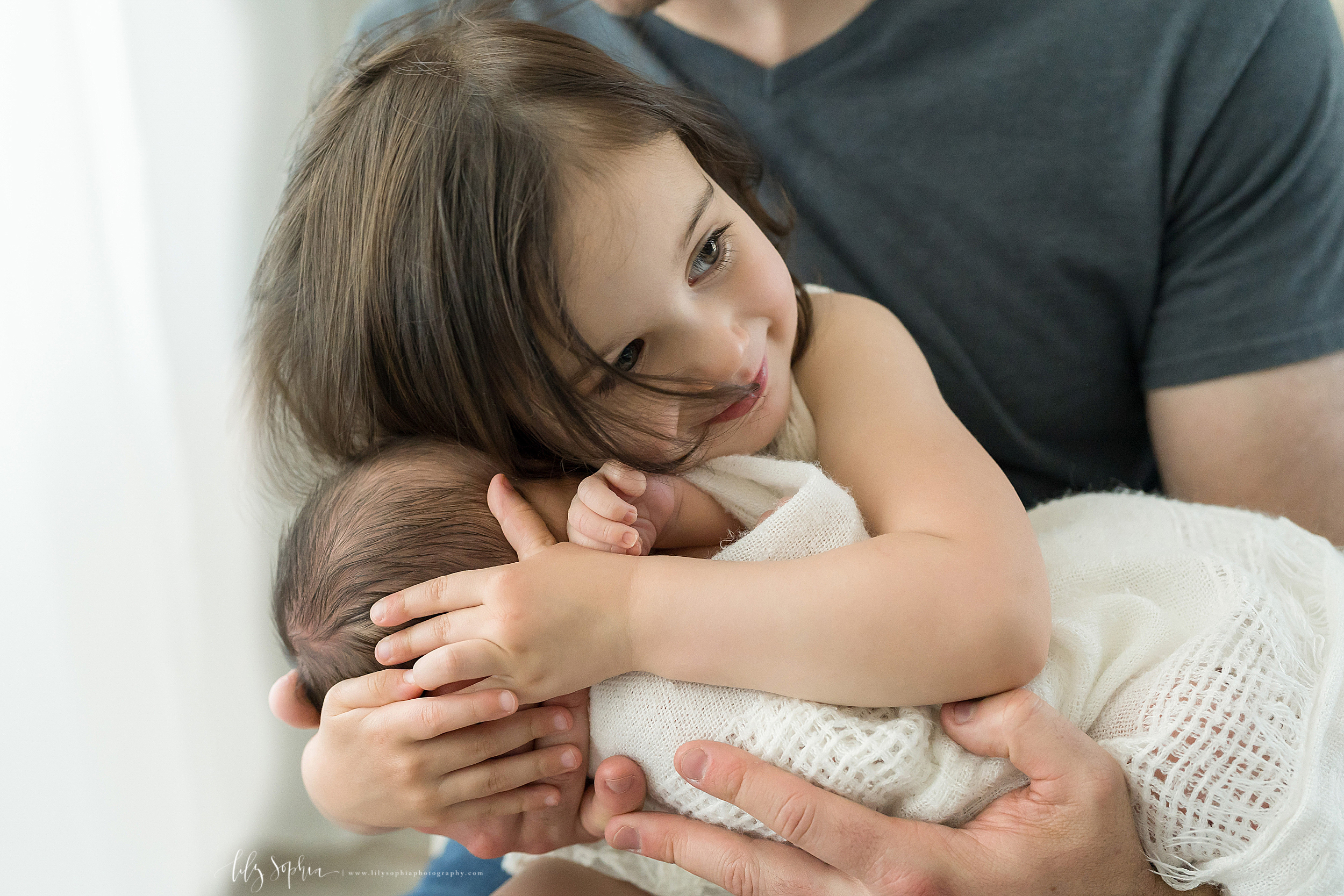  Dad is holding his infant son with brown hair wrapped in a white blanket on his lap. His toddler brunette daughter is giving her brother a giant hug while smiling. 