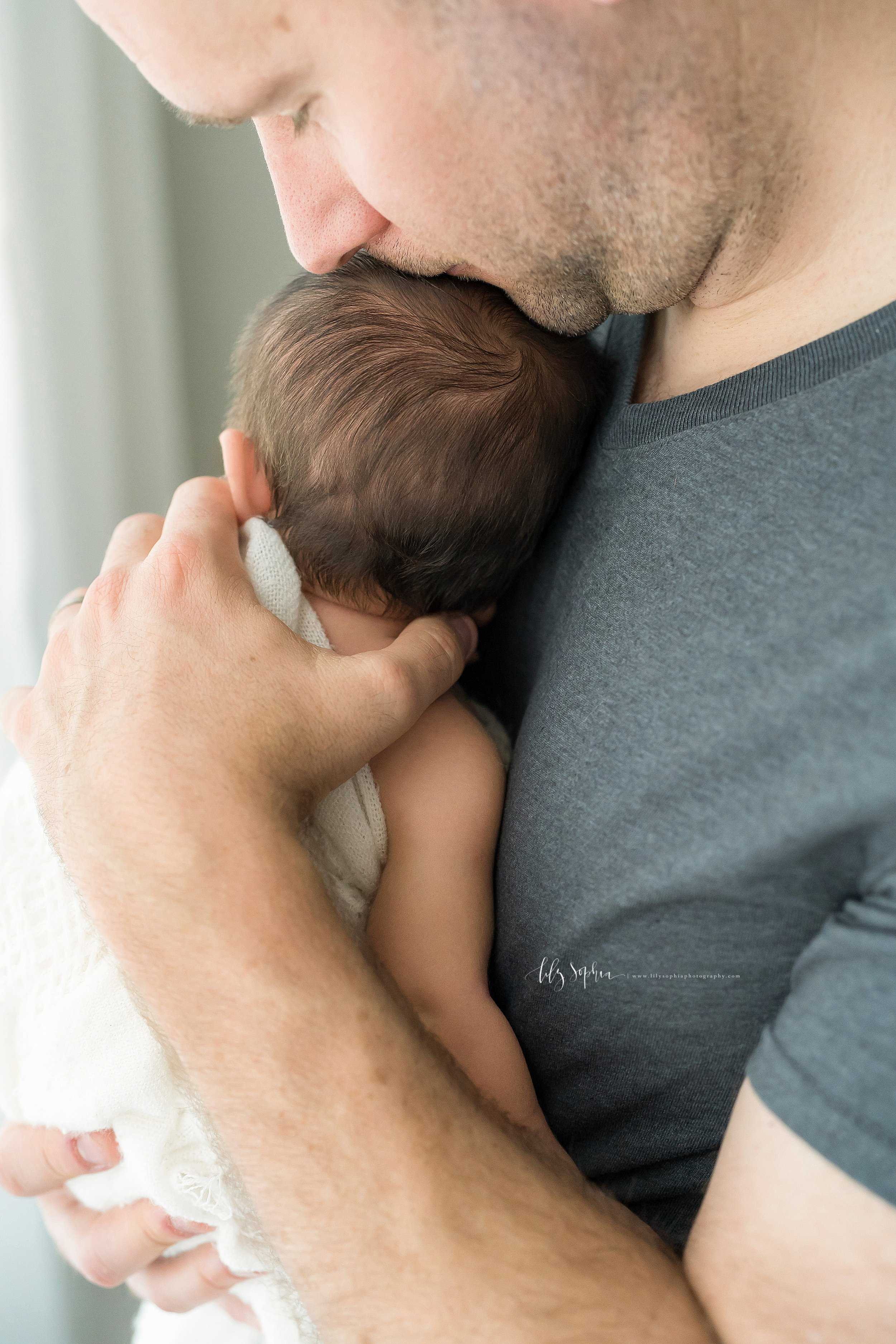  Dad wearing a gray T-shirt and holding his infant son against his chest stands in front of the window.  He kisses the top of his son’s brunette head. 