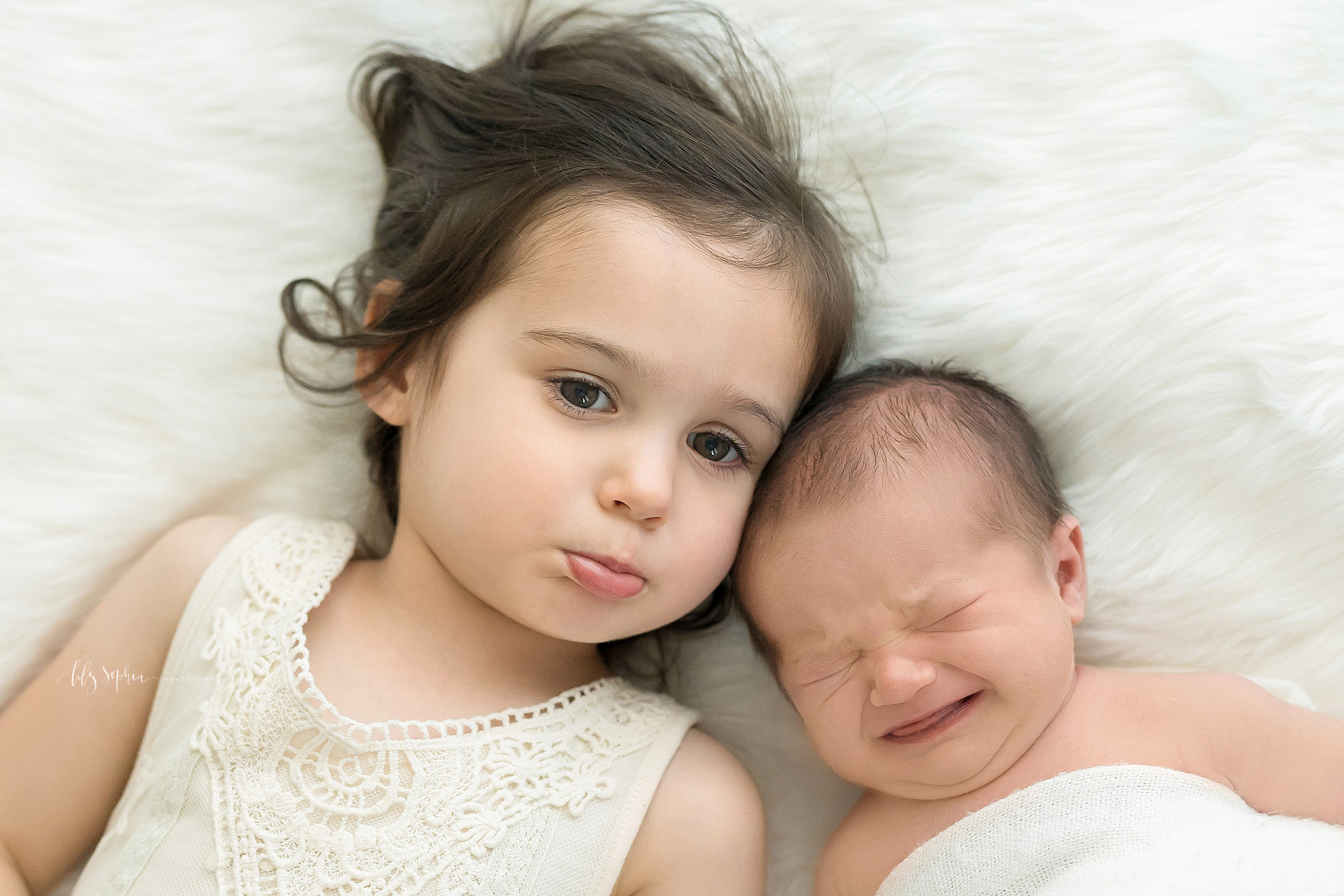  Siblings are lying next to each other on a soft white blanket.  The infant boy with brown hair is crying.  The toddler girl wearing a sleeveless white lace dress is pouting because she is sad that her brother is not happy.  