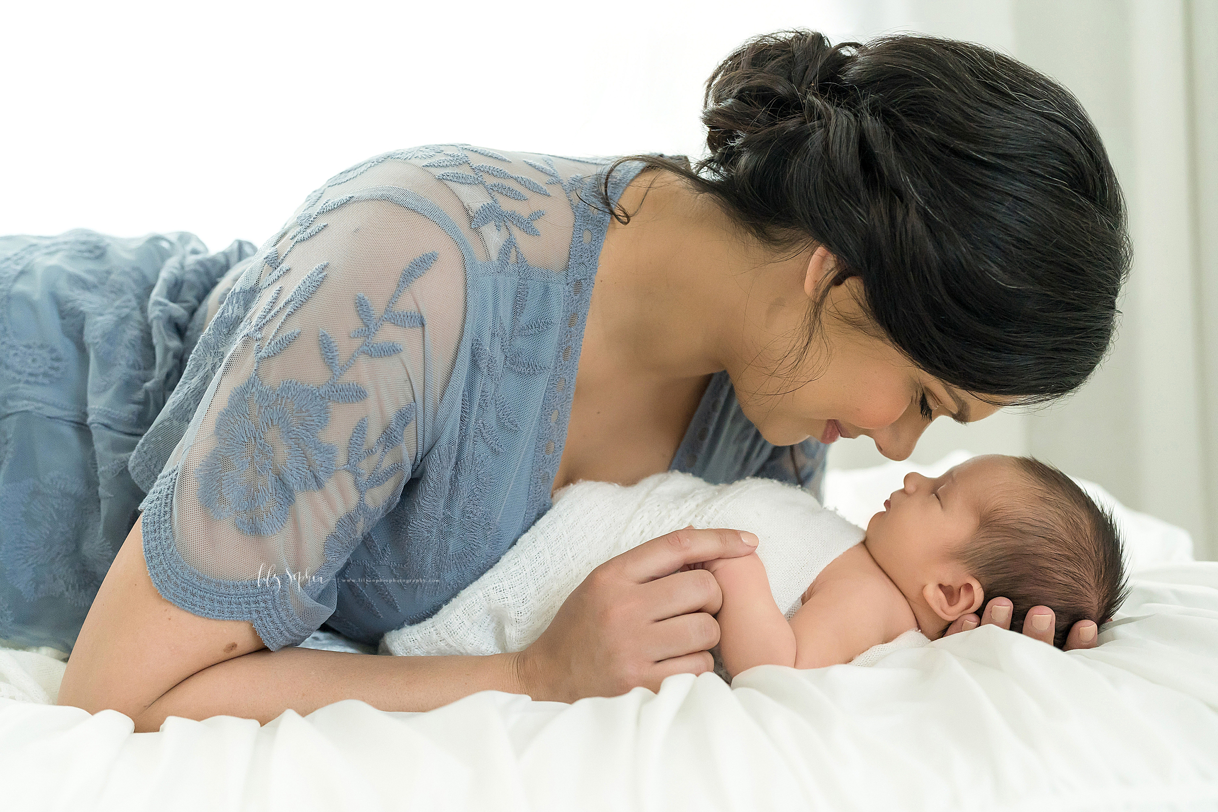  Brunette mother dressed in a blue lace dress is lying on a bed holding the hand of her infant son.  Her infant son is wrapped in a white blanket lying on his back sound asleep.  Mom is lovingly staring her infant son. 
