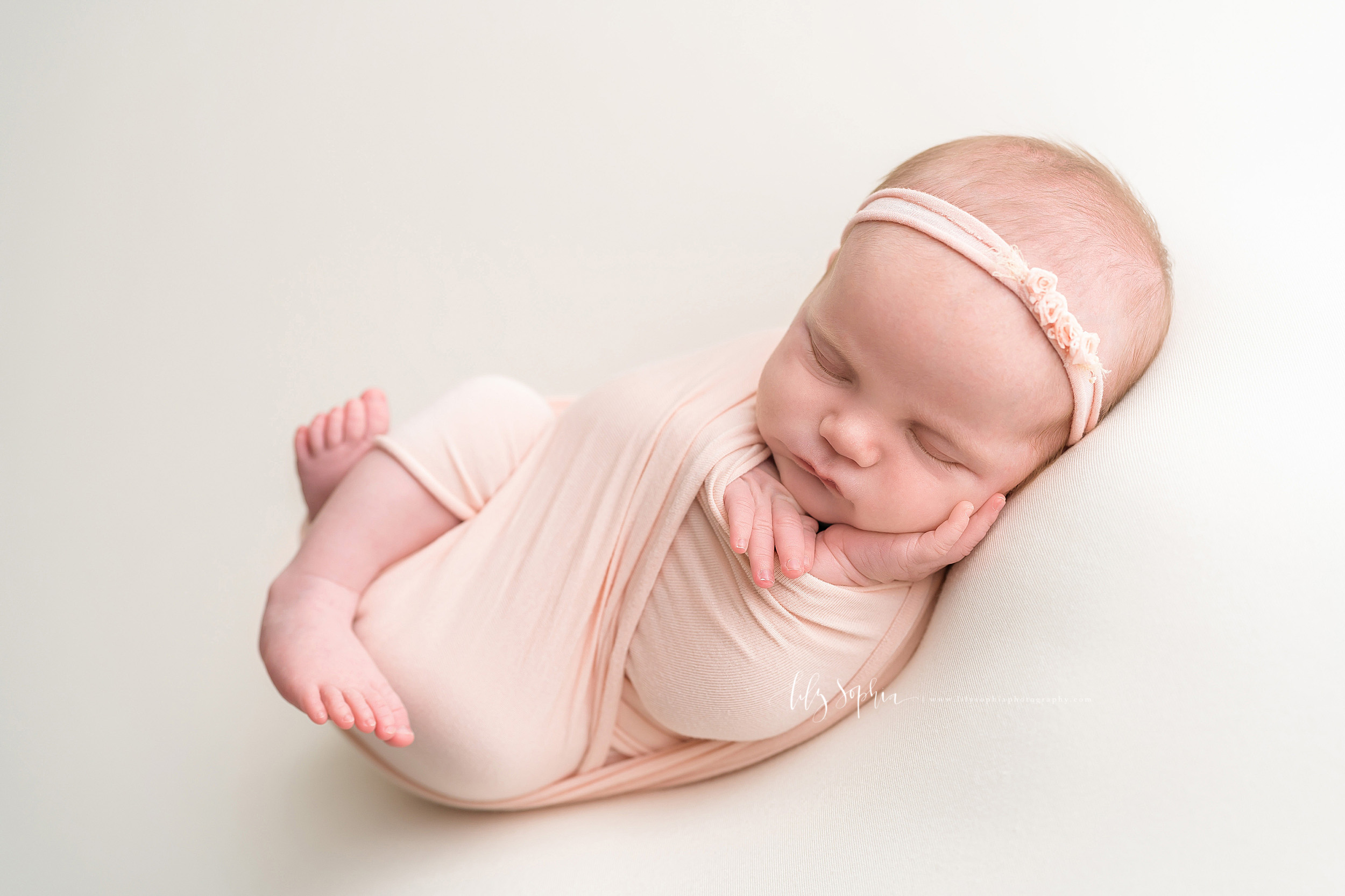  Image of a sleeping infant girl wrapped in a knit pale pink swaddle.  Her legs are bent and her feet are sticking out the bottom of the swaddle while her little hands are sticking out the top.  Her left hand is open and resting on her cheek.  She is