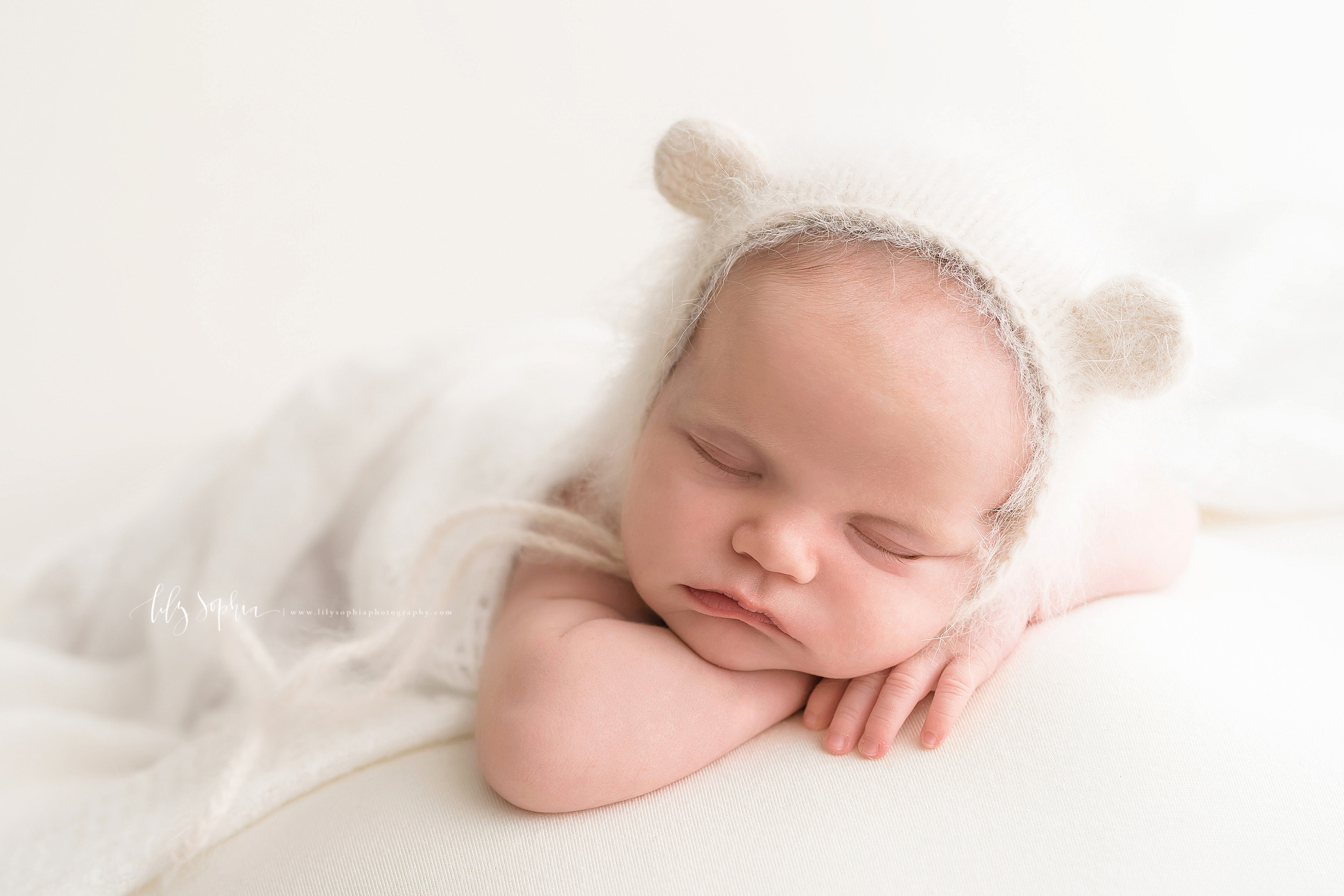  Photograph of an adorable sleeping infant girl in a natural light studio.  The infant is laying on her stomach with her arms under her head.  Her left arm is bent and she has her head resting on it.  The right hand and her little fingers are under h