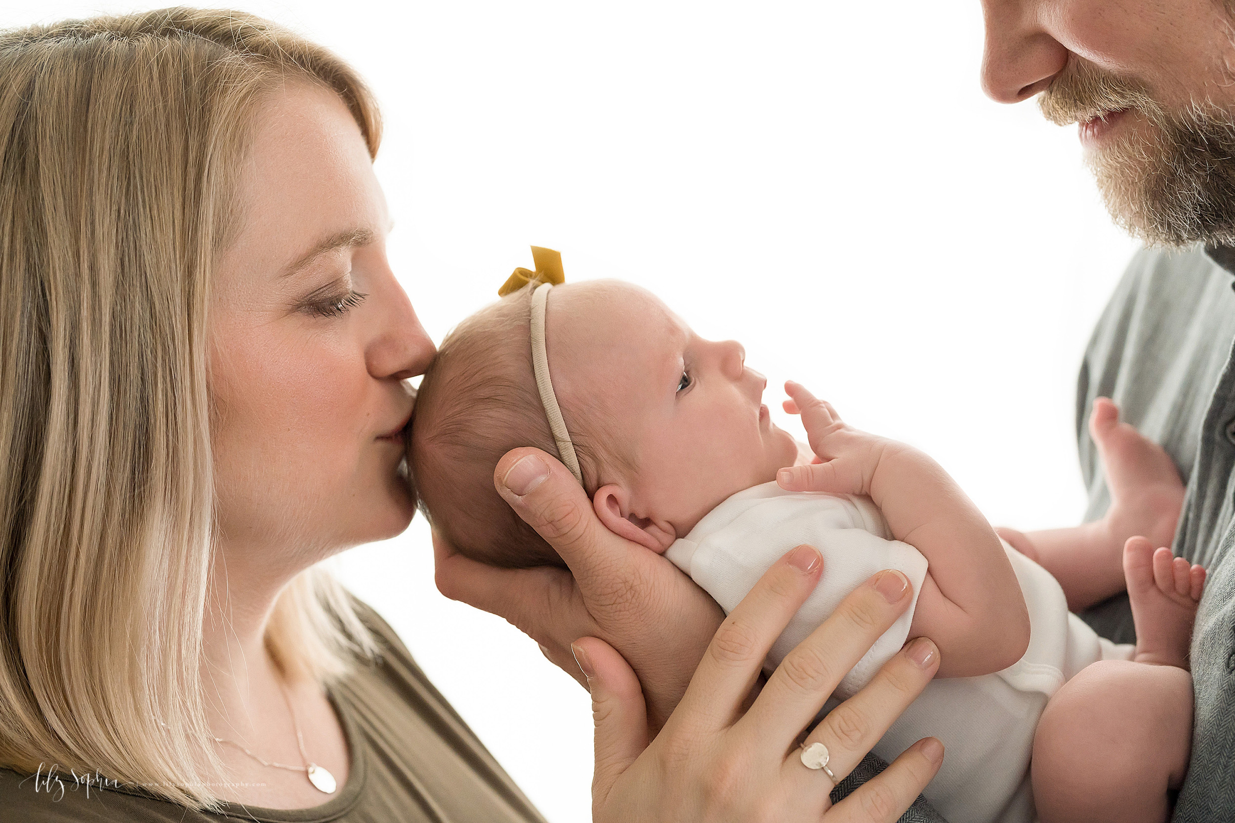  Image of a family of three in natural light.  The long blonde haired mom is facing her husband and kissing the top her her infant daughter’s head.  The father is facing his wife and cradling his infant daughter’s head in his hands.  Mom has her hand