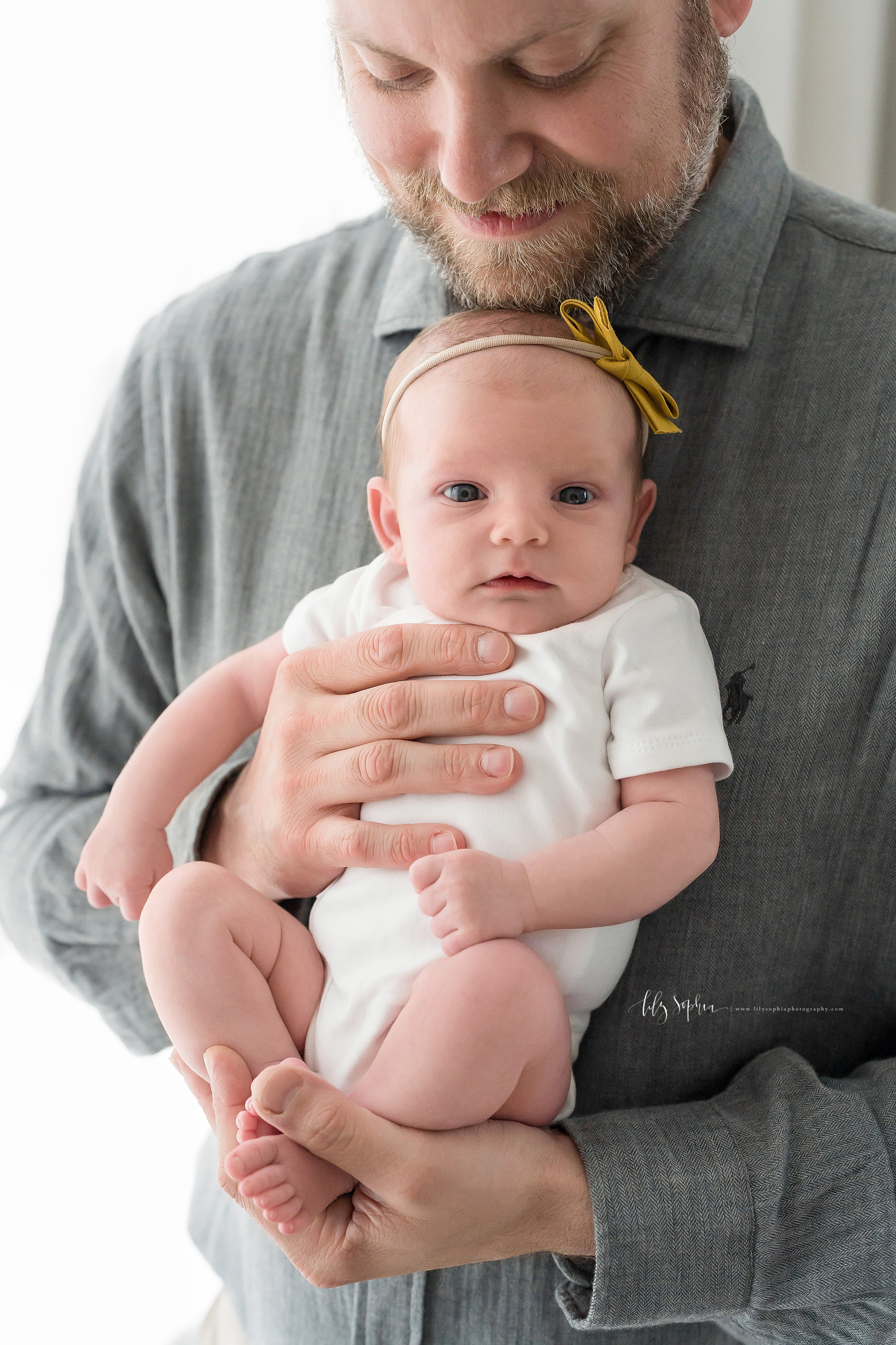  Image of a father holding his infant daughter in a studio in Atlanta. The bearded smiling father is wearing a long sleeved gray chambray shirt and is holding his infant daughter’s back against his chest while resting his chin on his daughter’s head 