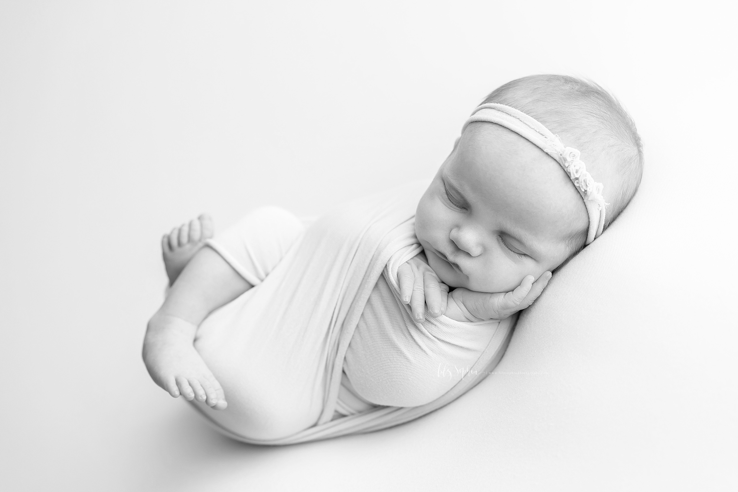  Black and white image of a sleeping infant girl wrapped in a knit  swaddle. Her legs are bent and her feet are sticking out the bottom of the swaddle while her little hands are sticking out the top. Her left hand is open and resting on her cheek. Sh