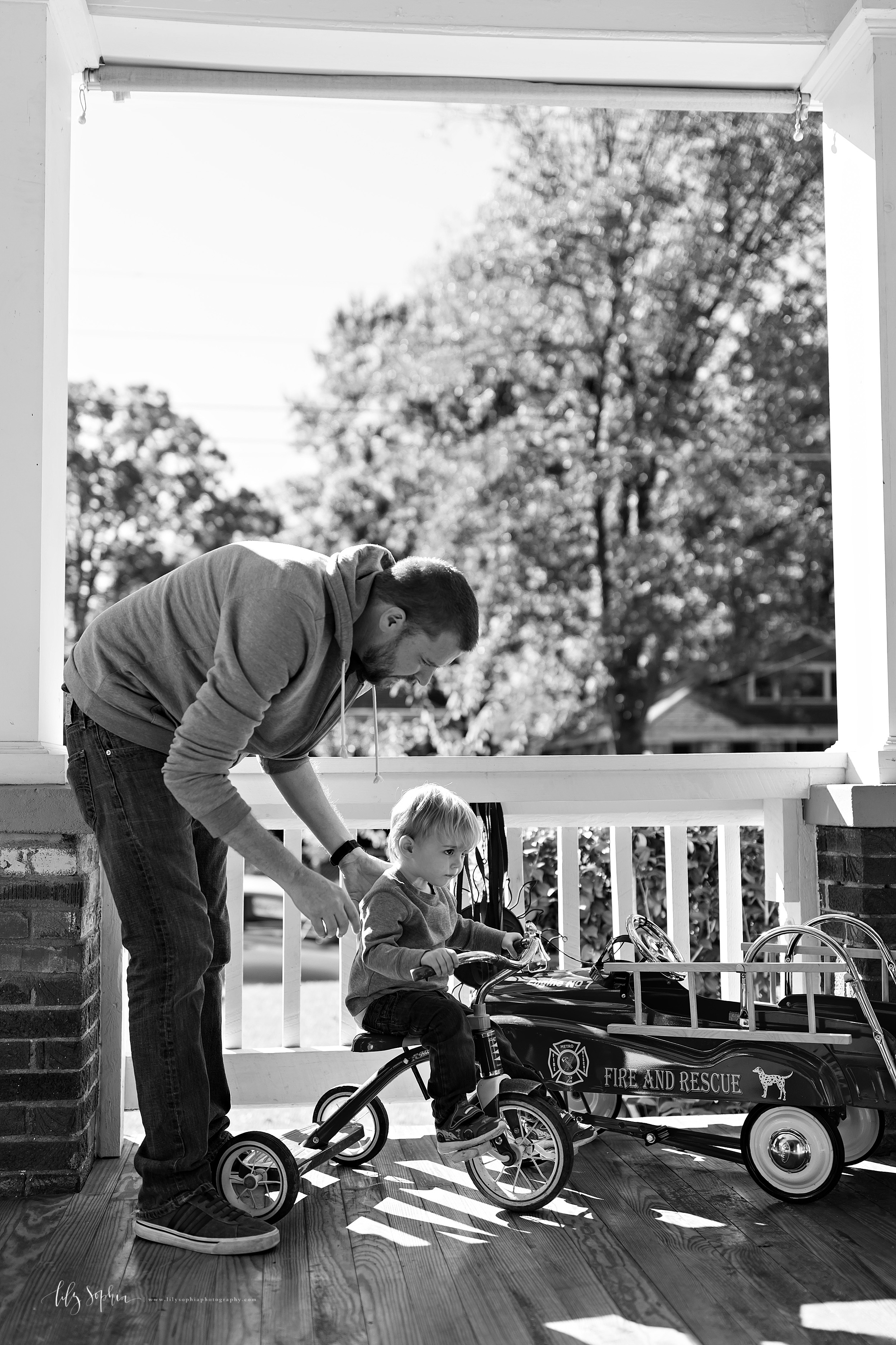  Black and white lifestyle photo of dad and his toddler son on their porch in Adair Park.  Dad is wearing a hooded sweatshirt and jeans.  His son is wearing a long-sleeved henley and jeans.  The toddler is seated on his tricycle learning how to ride.