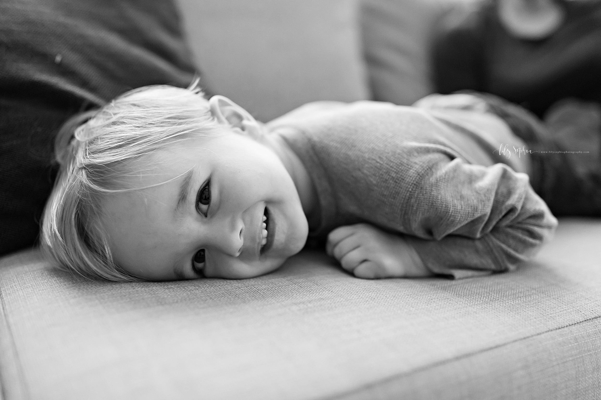  Black and white lifestyle photo of a smiling toddler boy as he lays face down on his couch at home.  He  is smiling as he looks over his left shoulder and has his left hand and arm under him. 