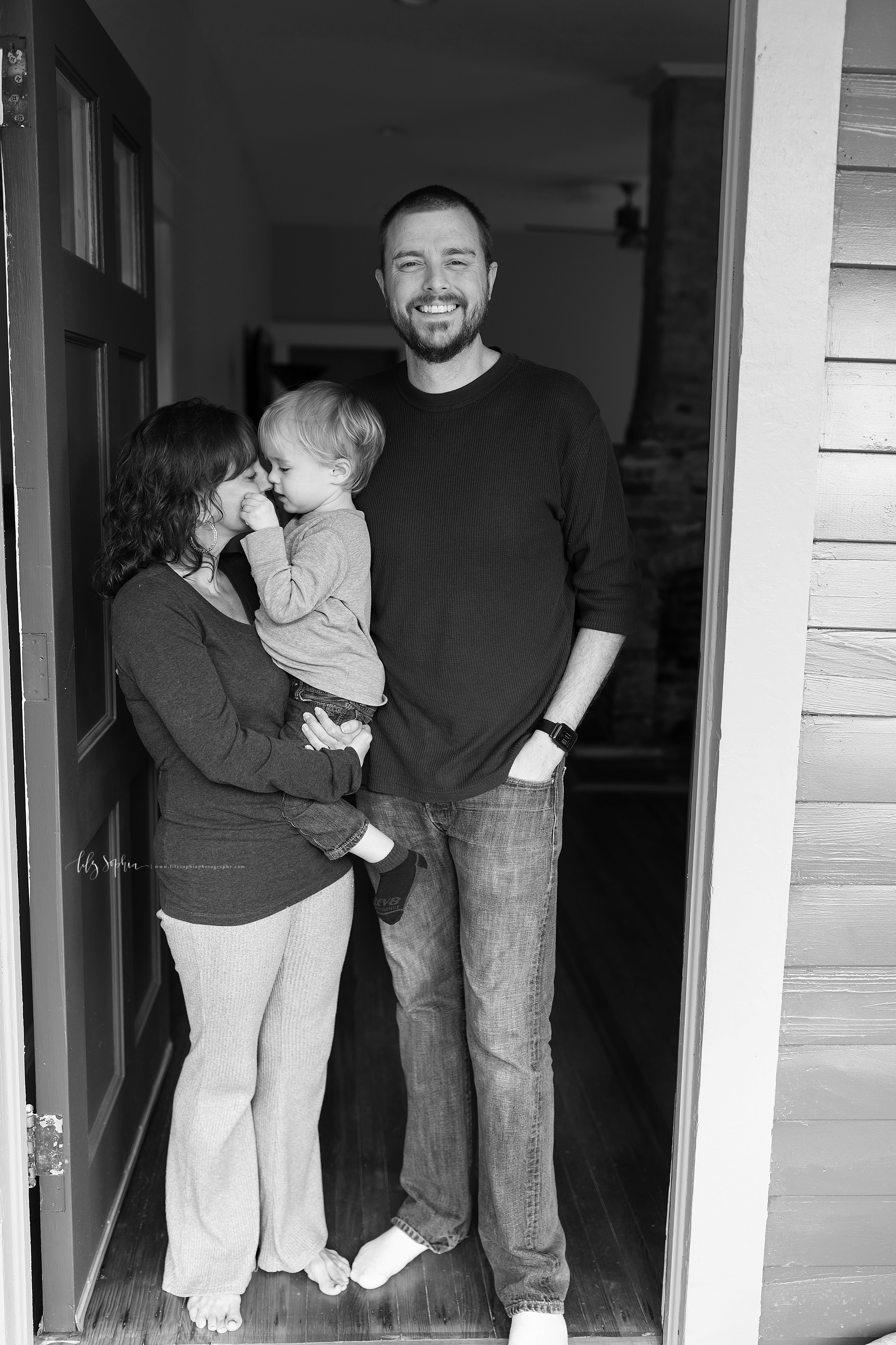  Black and white lifestyle photo of a family of three in Adair Park in Atlanta.  The family of three are standing in the doorway of their home.  Mom is holding her blond haired toddler son as he faces her and pinches her cheeks.  Dad is smiling as he