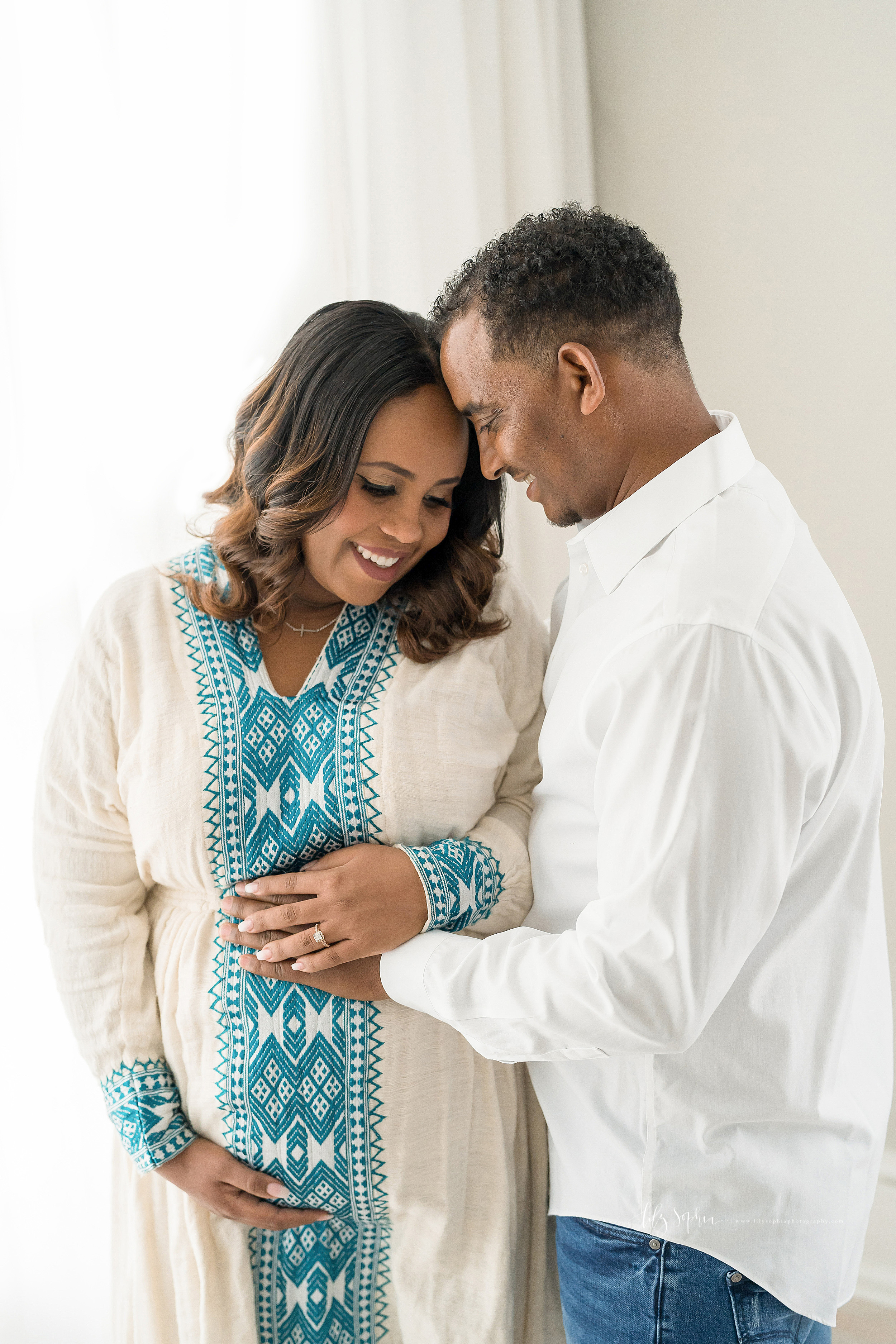  Maternity photo of Ethiopian couple in a natural light studio in Atlanta.  Mom is wearing a cream linen traditional Ethiopian dress with long sleeves and blue embroidery.  Dad is wearing a long sleeve white button down shirt with blue jeans.  Dad us