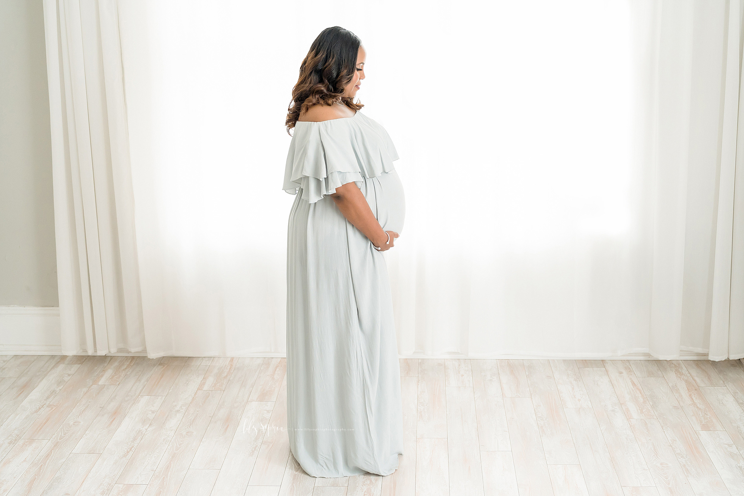  Maternity photo of an African American woman standing in front of a window in a natural light studio in Atlanta.  She is wearing a pale grey knit maternity dress that is off her shoulders.  It has a two-tiered large ruffle at the neckline.  Mom is h