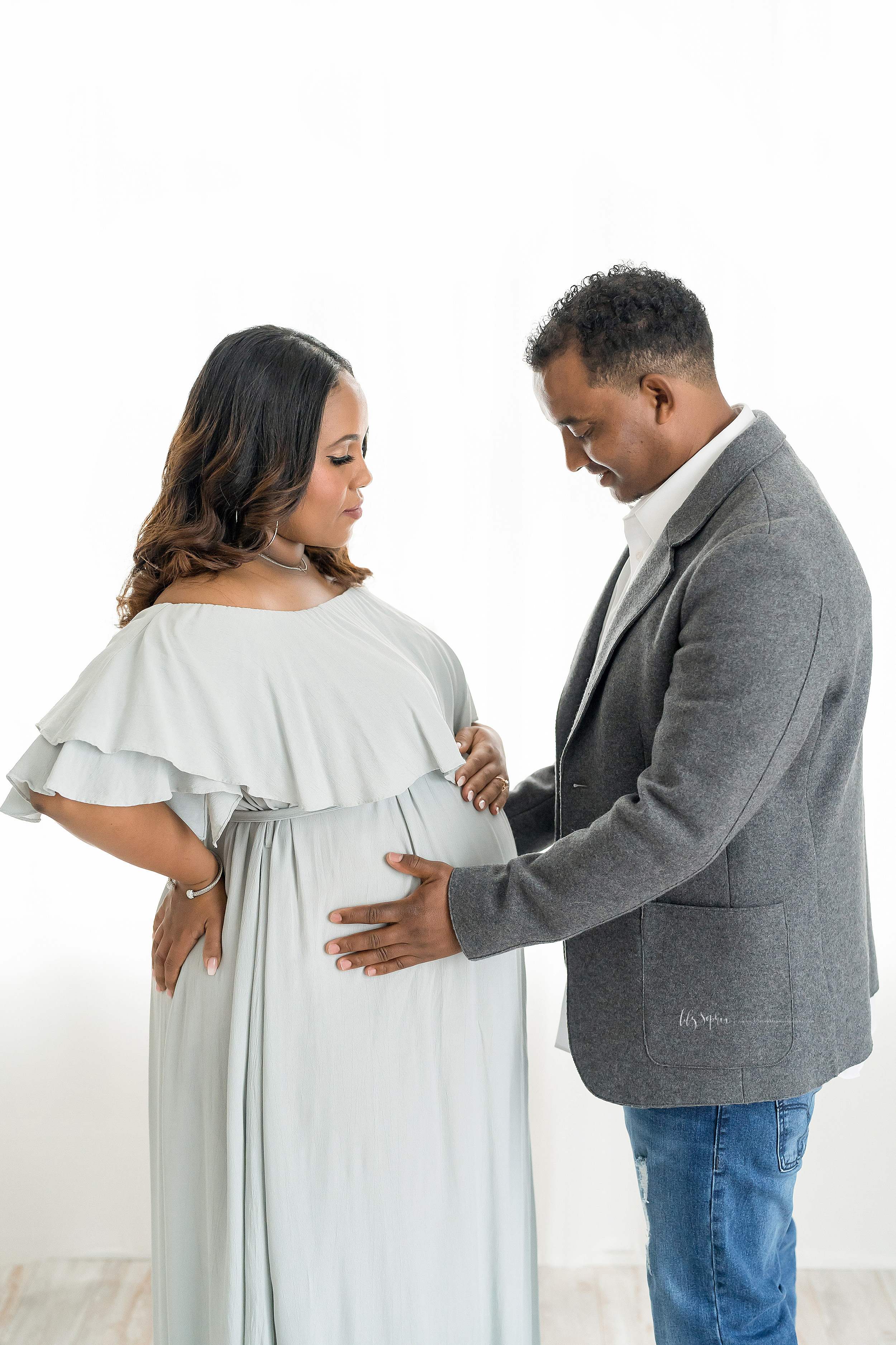  Maternity photograph of an African-American couple in a natural light studio in Atlanta. The husband and wife are standing and facing one another. The husband is on the right and is wearing a gray suit jacket over a white shirt and blue jeans. His w