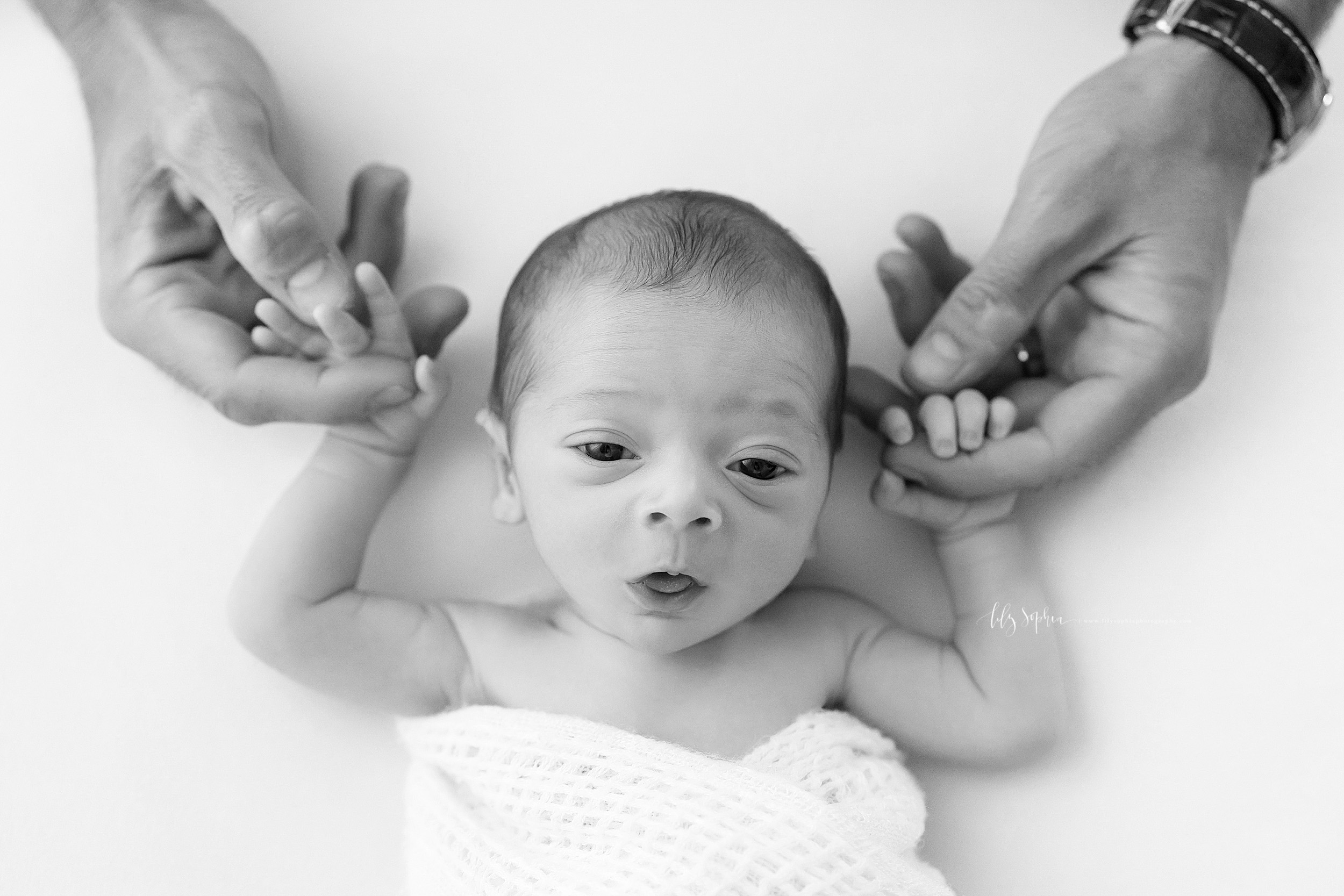  Black and white photo of Dad and his newborn son in Atlanta. The newborn boy is laying on his back with his eyes open. Dad’s strong hands can be seen as his son has his arms above his head and he is grasping his Dad’s pointer fingers.  The newborn’s