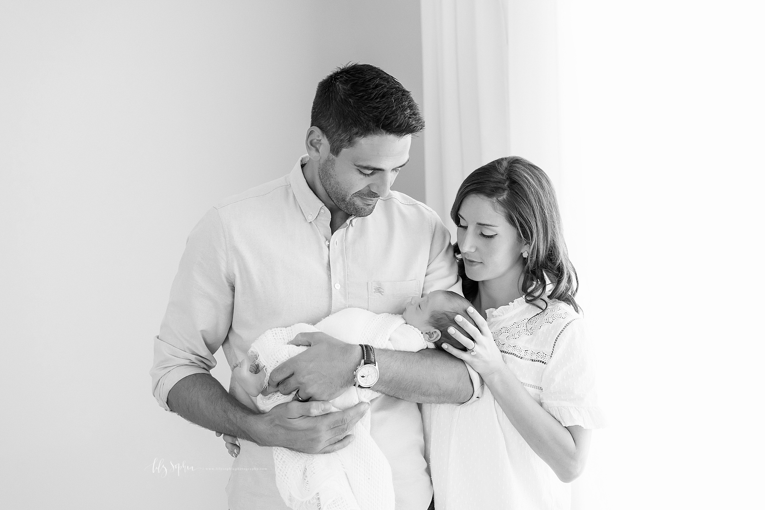  Photo in black and white of a family of three.  Dad is holding his newborn son in front of him.  The newborn’s head is in the crook of his right arm as he holds the baby’s body with his right hand and his left hand.  Mom is standing next to Dad.  Sh