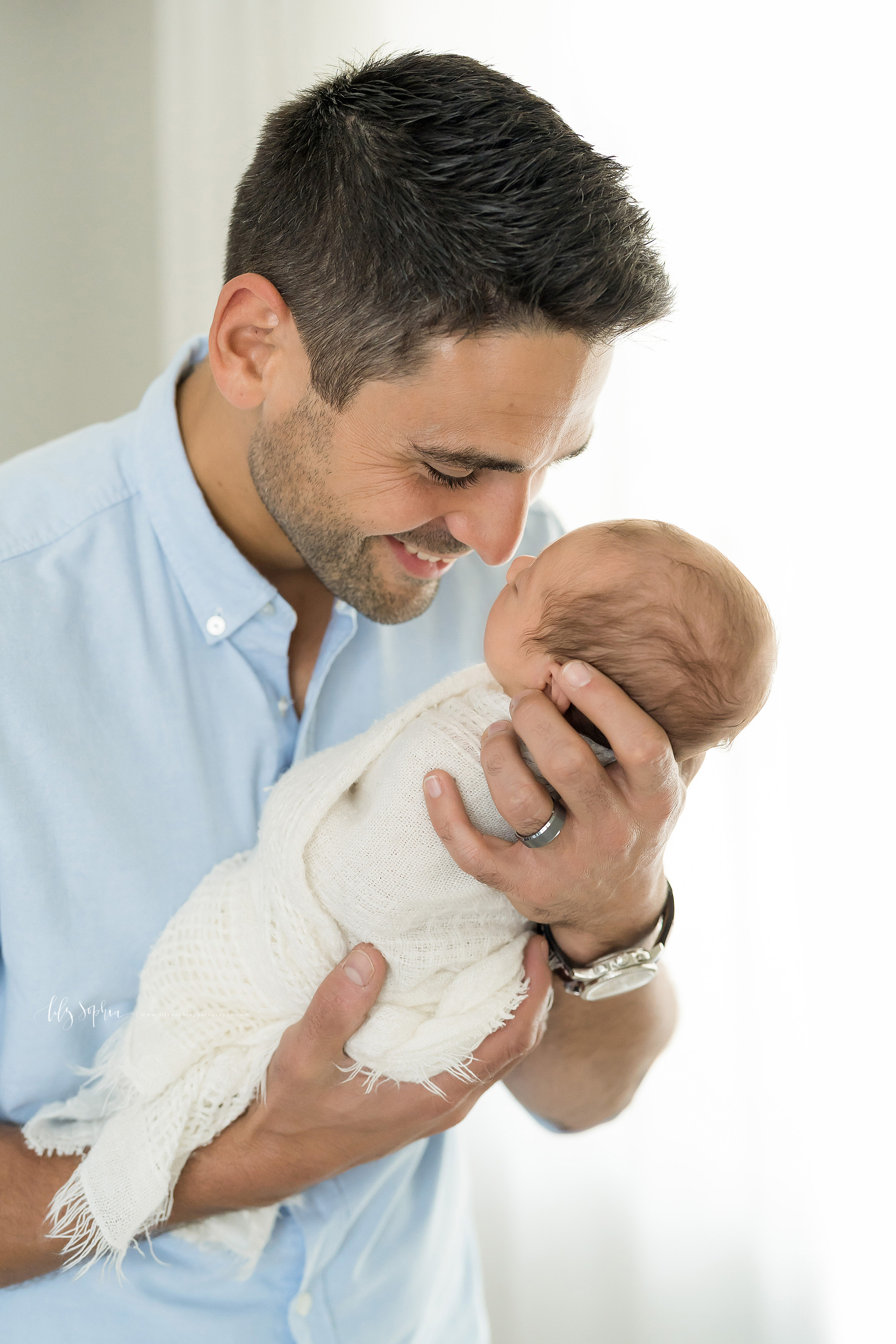  Photo of Dad and his newborn baby boy in Lily Sophia Photography Studio in Atlanta.  The brown haired Dad is wearing a light blue button down shirt.  His newborn son is wrapped in an off-white swaddle blanket with just his head peeking out.  Dad is 