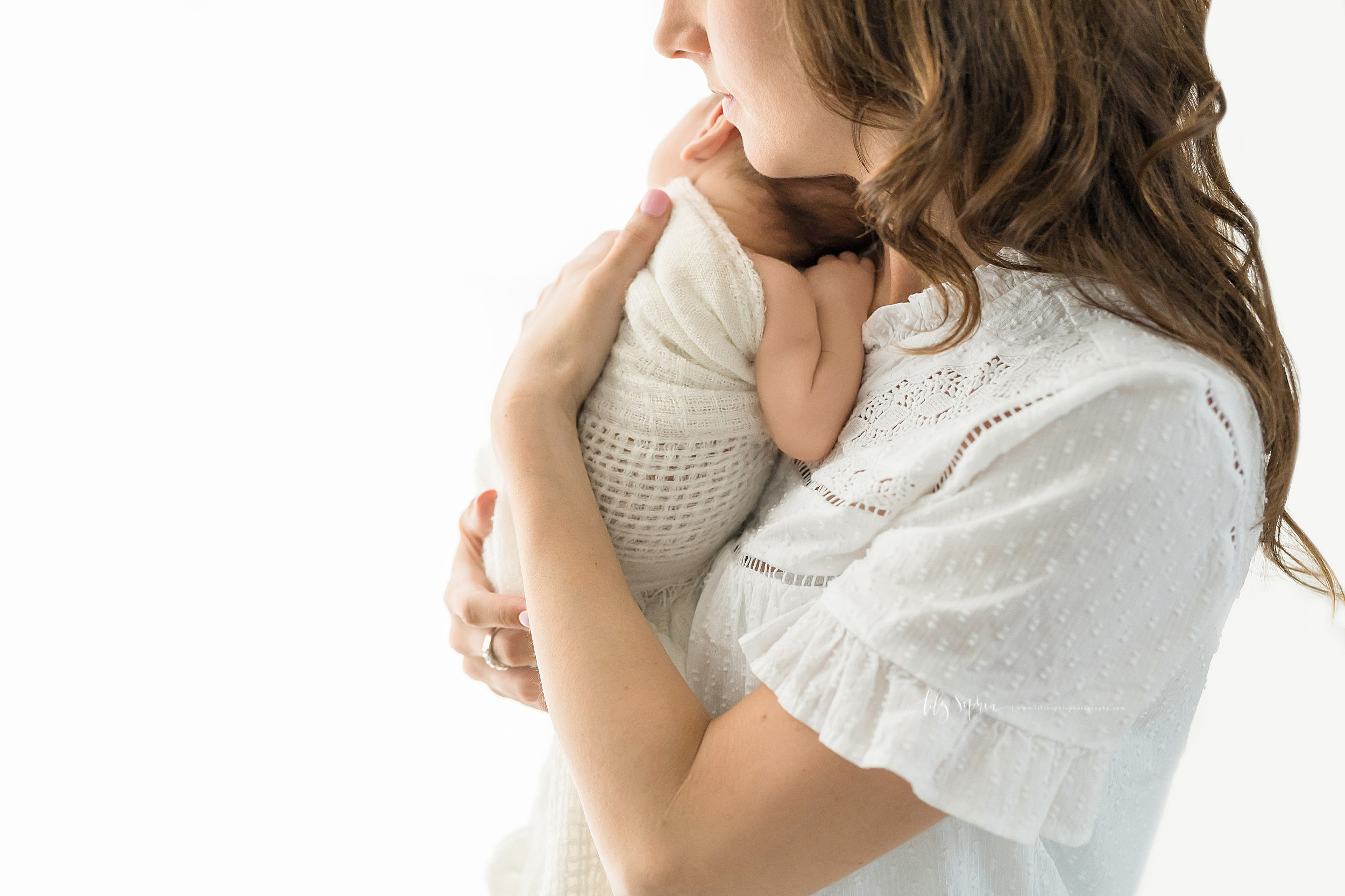  Photo of Mom and her newborn son in a natural light studio in Atlanta.  The newborn is wrapped in a  cream colored swaddle blanket.  Mom is wearing a white on white short sleeve cotton shirt.  She and the newborn are looking toward the right as Mom 