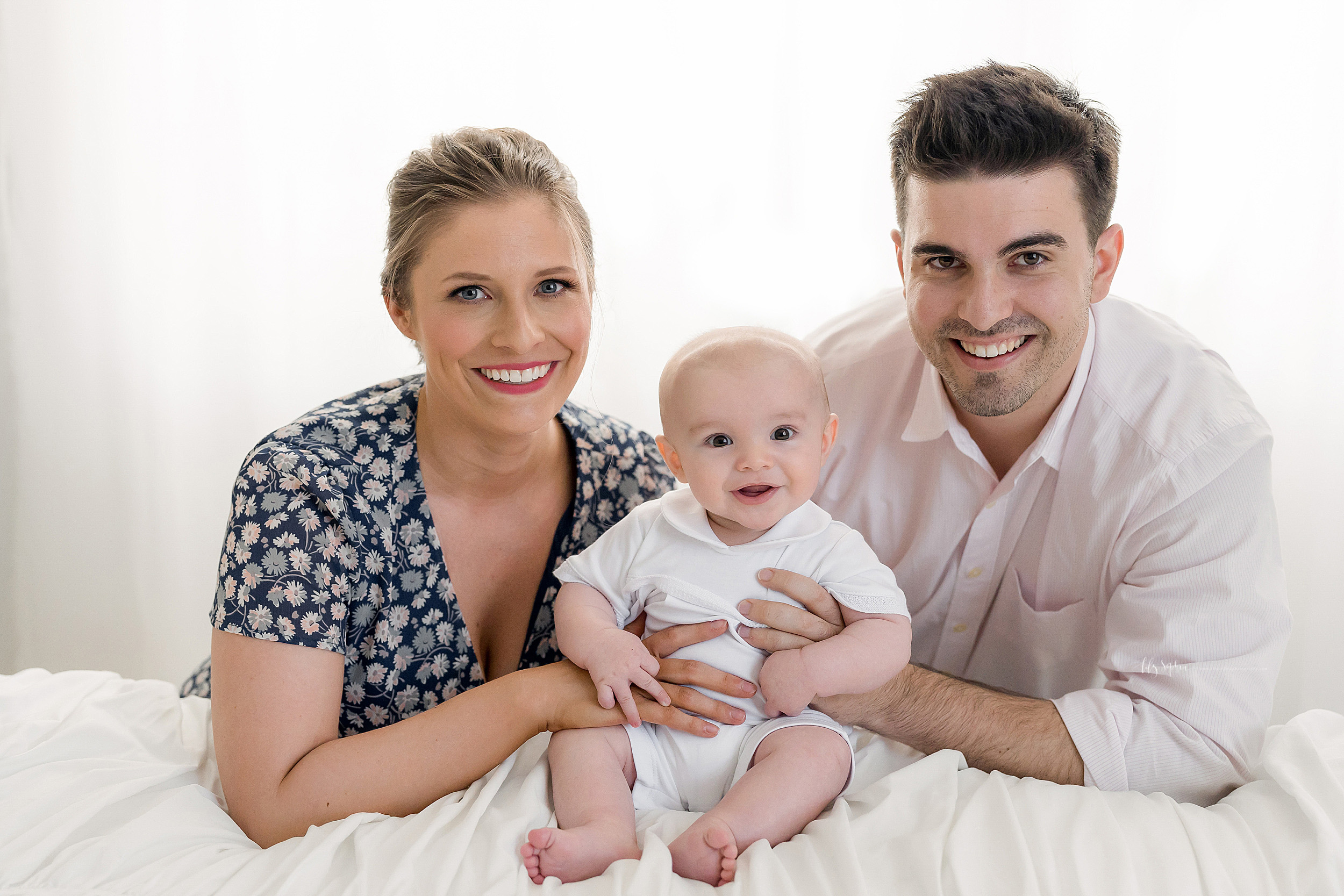 Photo of a happy family of three in a natural light Atlanta studio. Mom and Dad are kneeling on the floor behind a bed. Mom is on the left and Dad is on the right. Mom is holding her four month old son with her left hand as he sits and smiles on the bed and Dad is holding his son with his left hand, too. Blonde haired, blue-eyed Mom is wearing a V-necked navy blue dress with a daisy print. Brunette, brown-eyed Dad is wearing a pale pink long sleeve button down cotton shirt. The entire family is smiling.