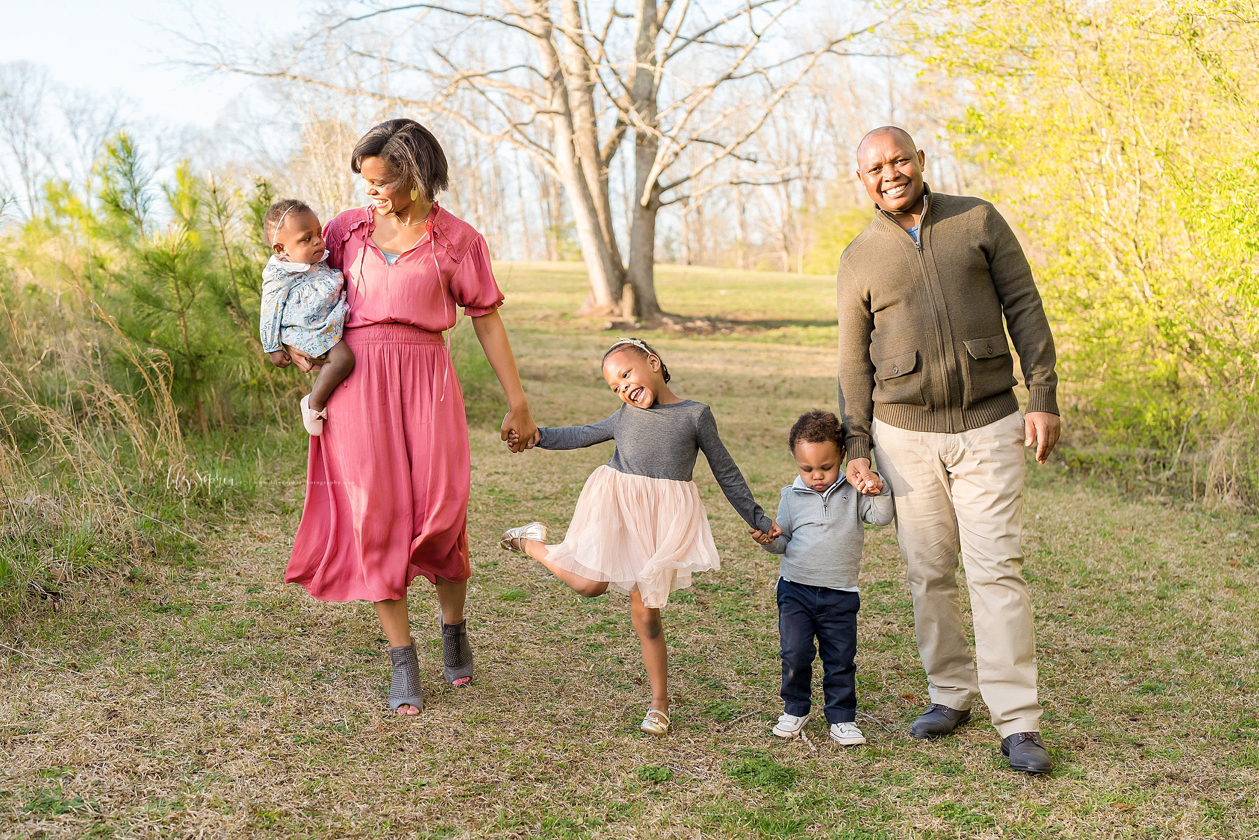  African-American family of five walk in an Atlanta field at sunset.  Mom is on the left holding her one year old daughter on her right hip.  Her other daughter is holding her mom’s left hand and her toddler brother’s hand.  Dad is on the right holdi