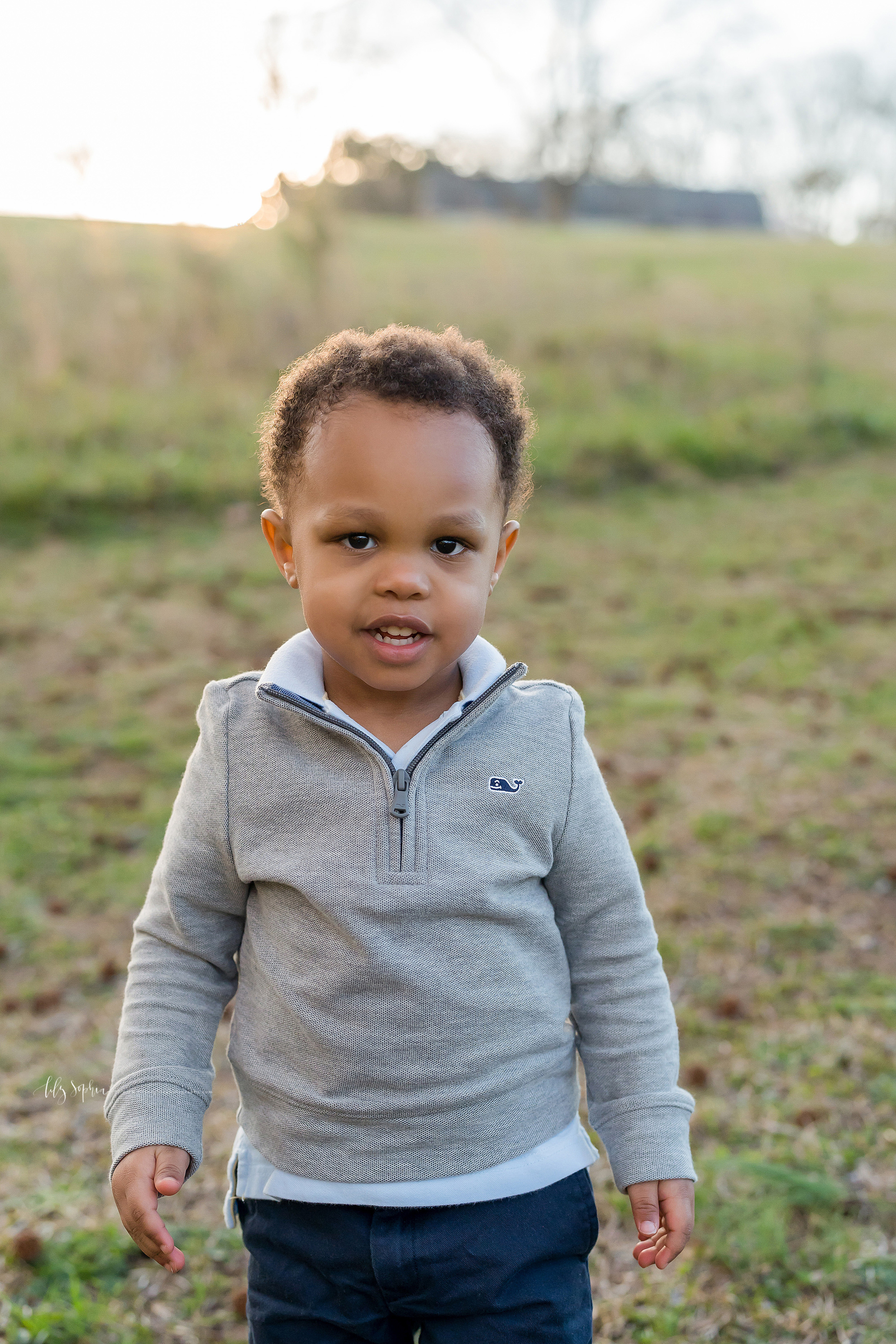  Photo of a toddler African-American boy at sunset in a field in Atlanta.  The curly haired, big brown-eyed boy is wearing a gray zippered sweatshirt over a white polo shirt and blue jeans.  He is standing in the field as the sun glows above his head