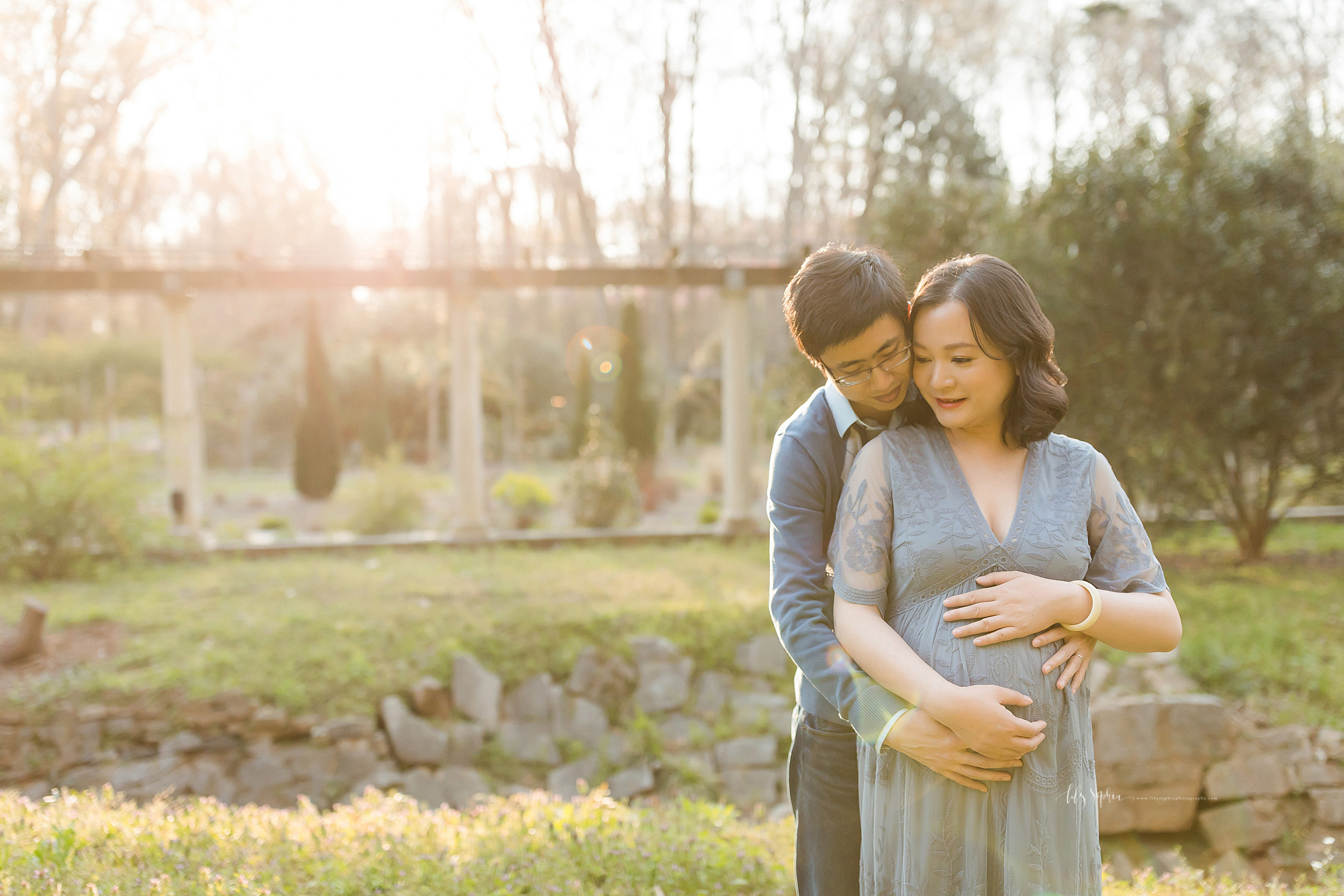  Pregnant Asian couple in at Atlanta garden for a photo shoot with Lily Sophia Photography at sunset. The husband is standing behind his wife with his right cheek against her left cheek. His wife is holding the top of her womb with her left hand. The