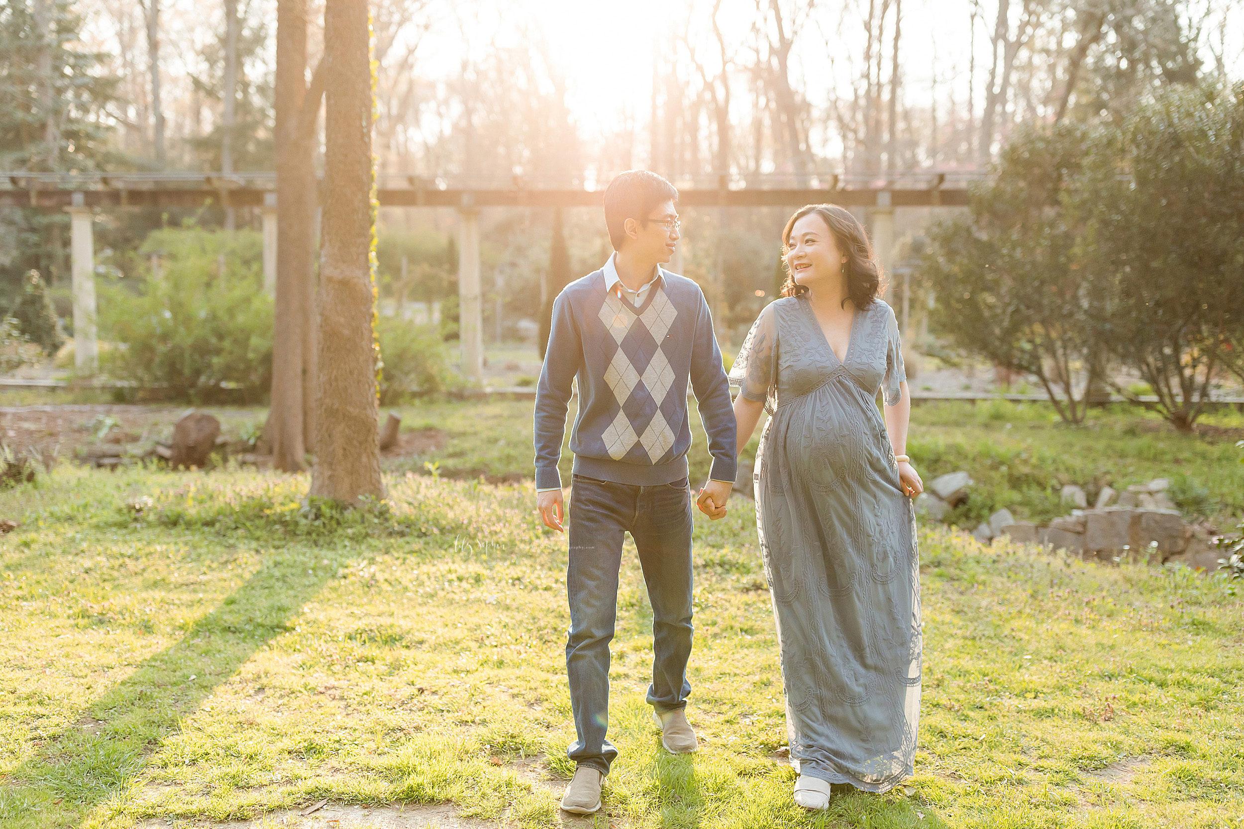  Maternity photo of an Asian couple walking in a garden at sunset in Atlanta. The husband is wearing a light blue shirt under a wedgewood blue pull over sweater with an argyle print on the front of the sweater and blue jeans. His wife is wearing a sh