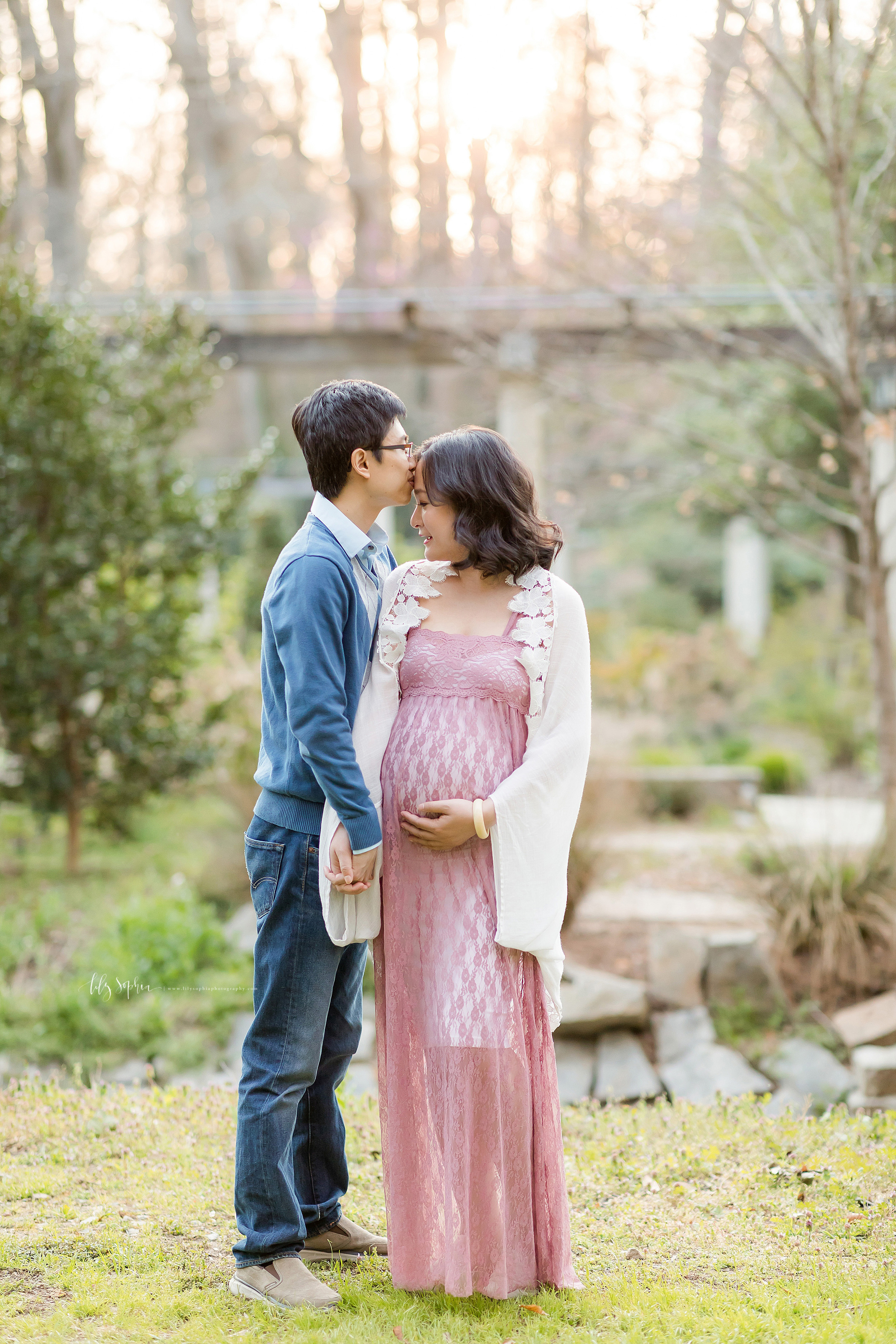  Maternity photo of an Asian woman and her husband taken by Lily Sophia Photography in an Atlanta garden at sunset.  The profile of the husband is seen as he holds his wife’s right hand with his left hand and kisses her forehead.  The wife is forward